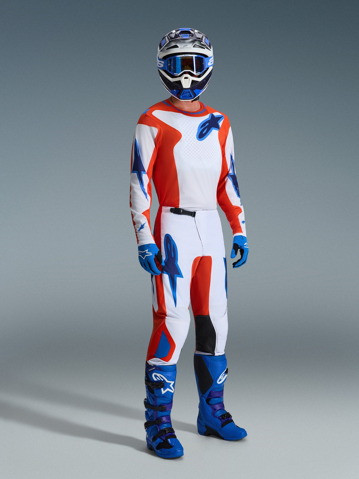 Motocross rider wearing the Alpinestars 2026 Fluid Grid Gear Set, white jersey and pants with orange and UCLA blue graphics, matching blue helmet and goggles, blue gloves, and bright blue racing boots, coordinated performance outfit for off-road riding.