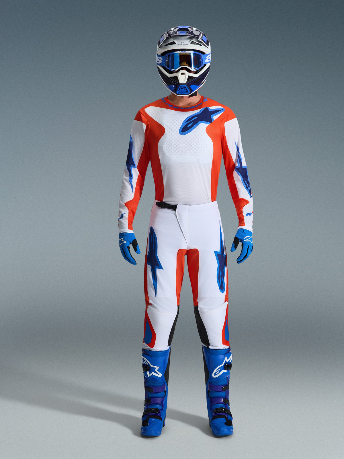 Pilote de motocross Alpinestars en tenue de course complète, maillot Fluid Grid 2026 Orange et UCLA Blue, pantalon blanc assorti avec accents orange et bleus, casque bleu et blanc avec lunettes bleues, bottes tout-terrain bleues et violettes, gants bleus, équipement de motocross coordonné