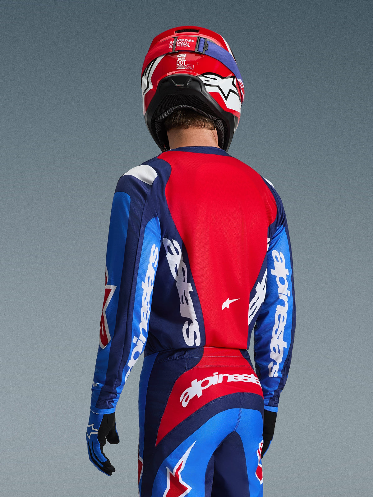 Rear view of a motocross rider wearing the Alpinestars 2026 Fluid Grid Jersey in Blue and Red, featuring bold white branding on the sleeves and back, paired with matching motocross pants and a red helmet for a complete off-road racing look