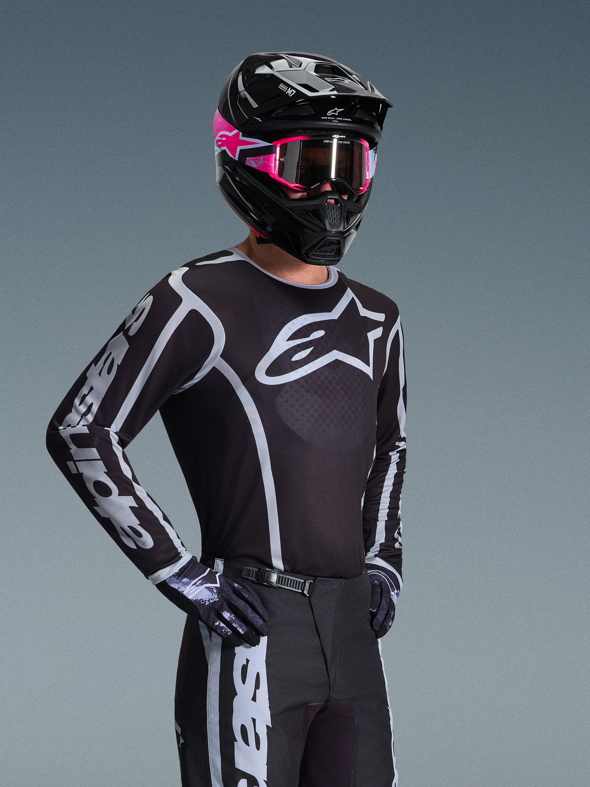 Pilote de motocross portant le maillot Alpinestars Fluid Apex 2026, noir et gris avec un grand logo blanc sur la poitrine et des graphismes géométriques sur les manches, associé à un pantalon noir, des gants noirs et un casque noir avec des touches roses et des lunettes roses