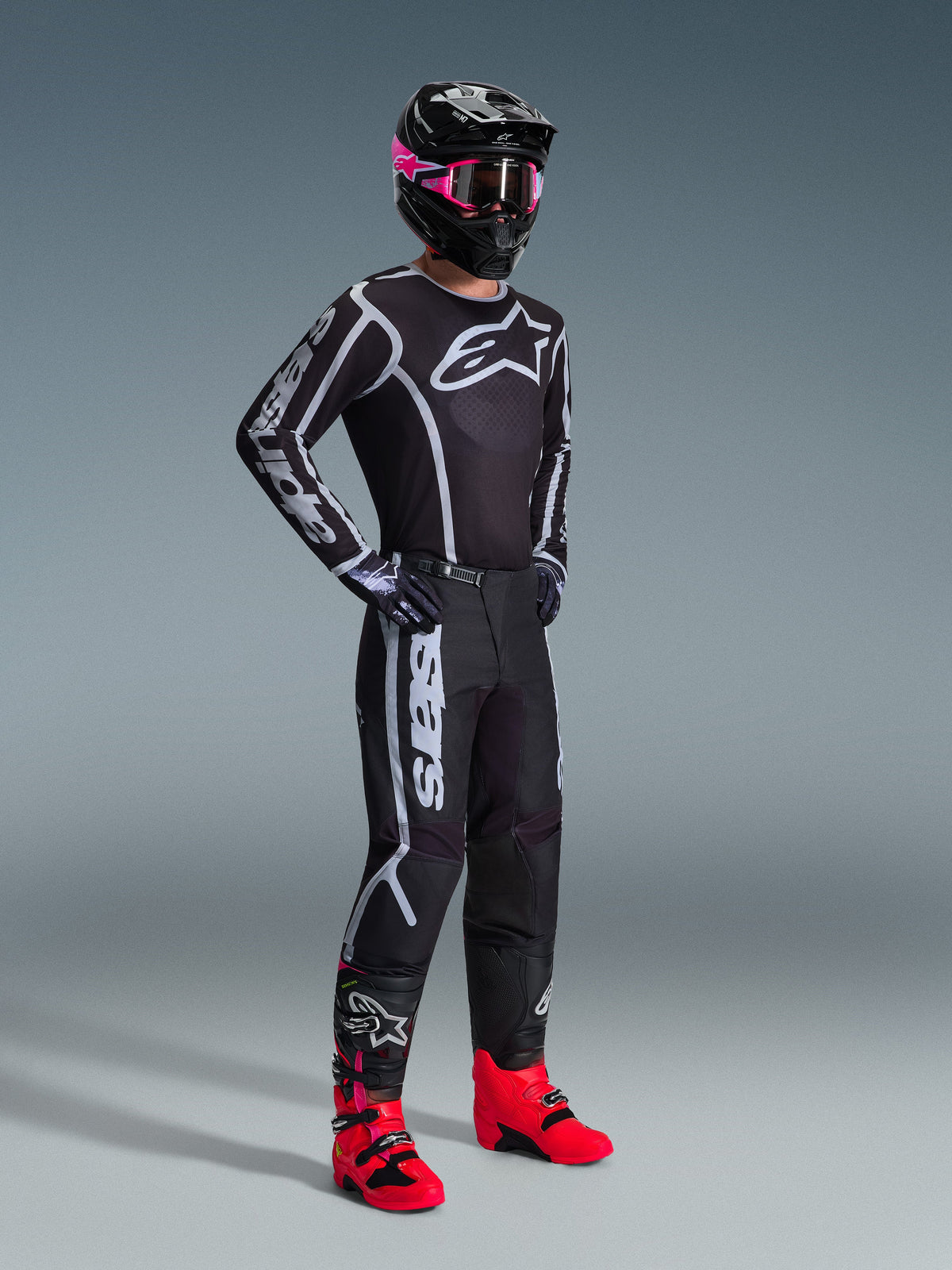 Pilote de motocross portant le maillot 2026 Fluid Apex, noir et gris, associé à un pantalon assorti, un casque noir avec des lunettes roses et des bottes rouge vif, avec le logo Alpinestars blanc sur la poitrine et les manches, tenue de course tout-terrain professionnelle coordonnée.