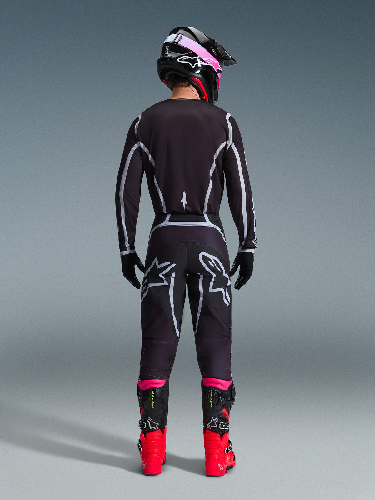 Pilote de motocross vu de dos portant le maillot Alpinestars Fluid Apex 2026 et le pantalon assorti Noir/Gris, avec des lignes géométriques blanches et des logos étoiles sur l'assise, accompagné d'un casque noir et rose et de bottes de course rouges et noires.