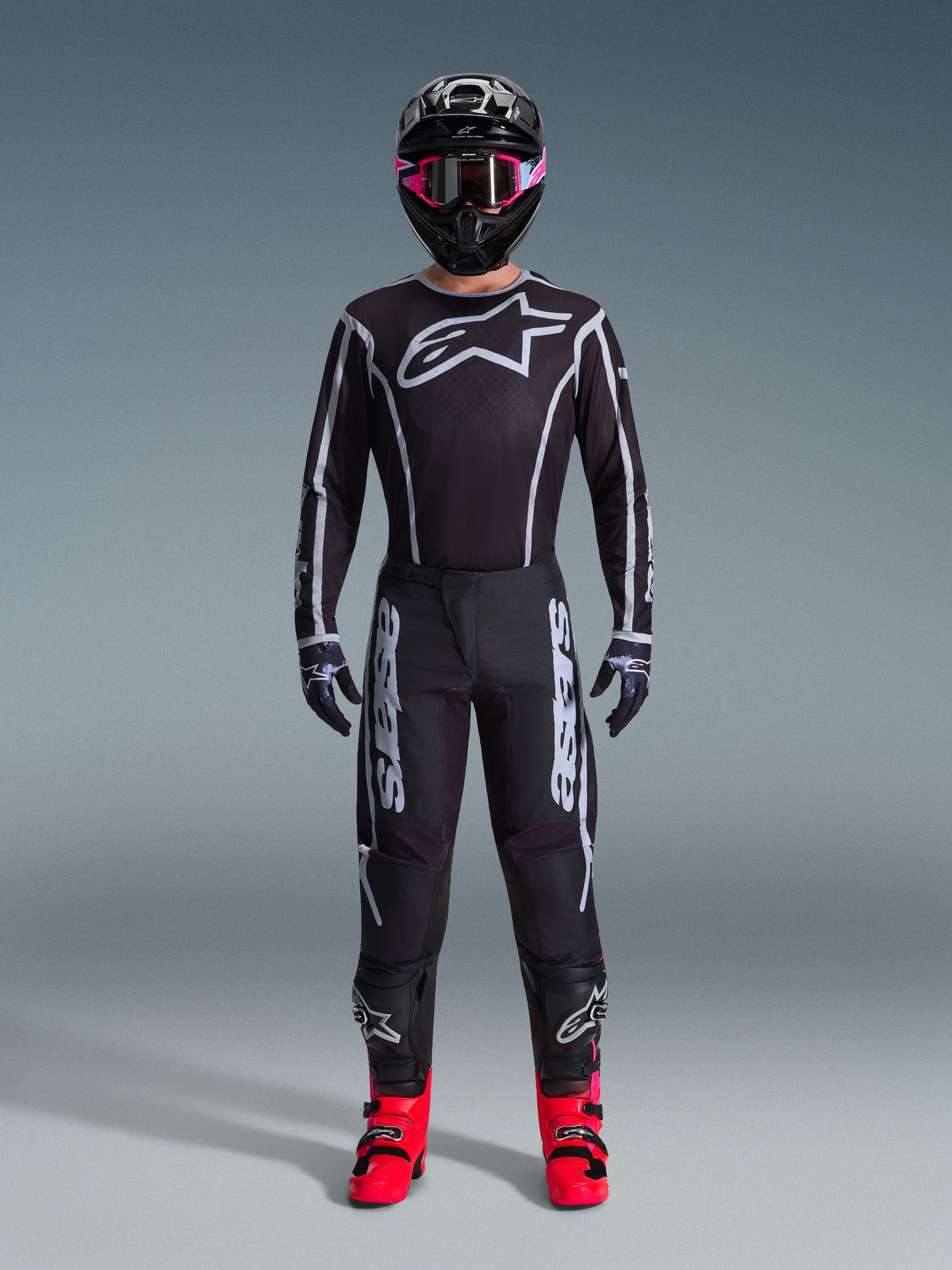 Pilote de motocross portant le maillot Alpinestars Fluid Apex 2026, noir et gris, grand logo Astars blanc sur la poitrine, lignes géométriques blanches, pantalon noir assorti, casque noir avec lunettes roses et bottes rose vif, tenue de course de motocross complète