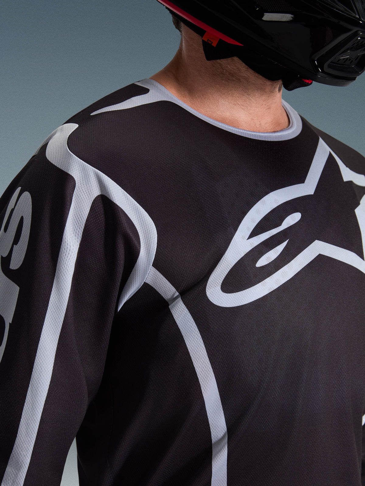 Gros plan d'un pilote portant le maillot Alpinestars 2026 Fluid Apex noir et gris, mettant en avant le grand logo Astars blanc sur la poitrine et la construction technique en mesh, avec un casque de motocross noir.