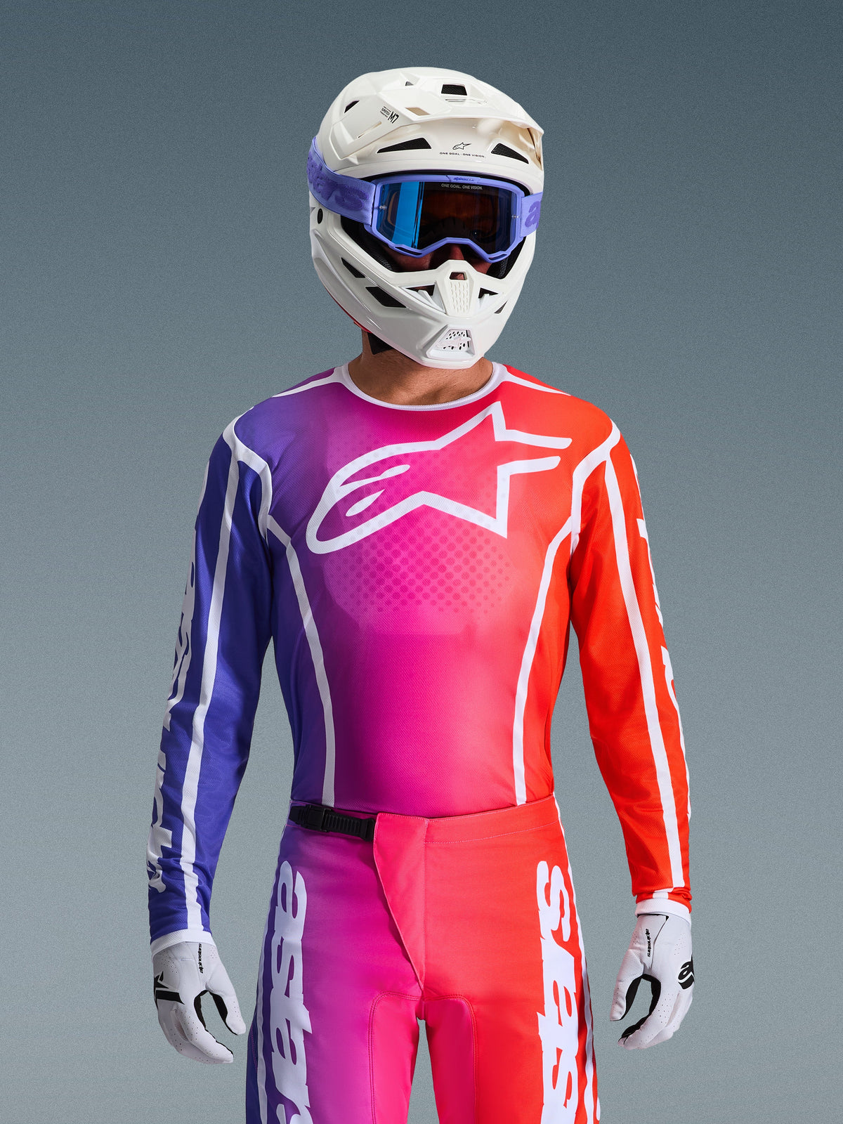Pilote de motocross portant le maillot Alpinestars Fluid Apex 2026, design en dégradé éclatant de violet, rose et orange avec un grand logo blanc sur la poitrine, assorti à un casque blanc, des lunettes violettes et des gants blancs pour une tenue de course tout-terrain complète.