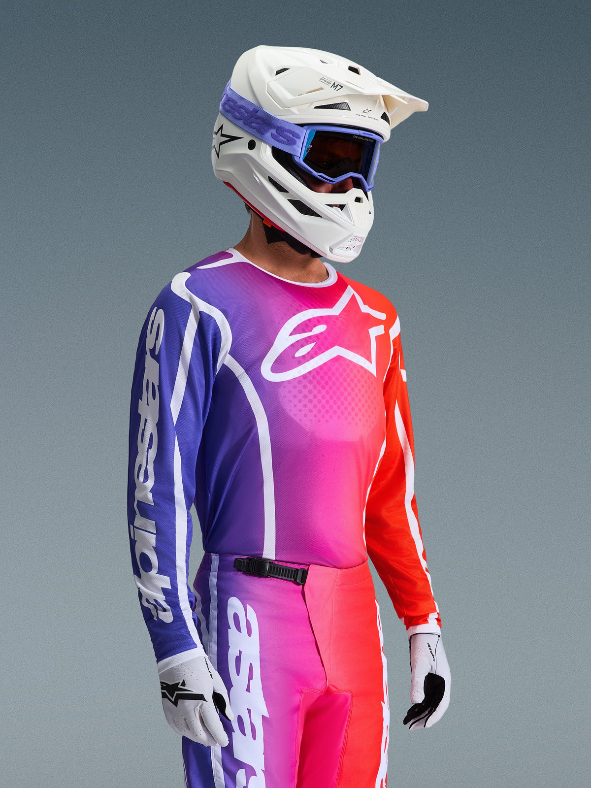 Maillot Alpinestars 2026 Fluid Apex, maillot de motocross, dégradé éclatant de violet, rose et orange, porté avec un pantalon assorti, casque blanc avec des lunettes bleues et des gants blancs, design athlétique pour la performance et la mobilité en tout-terrain