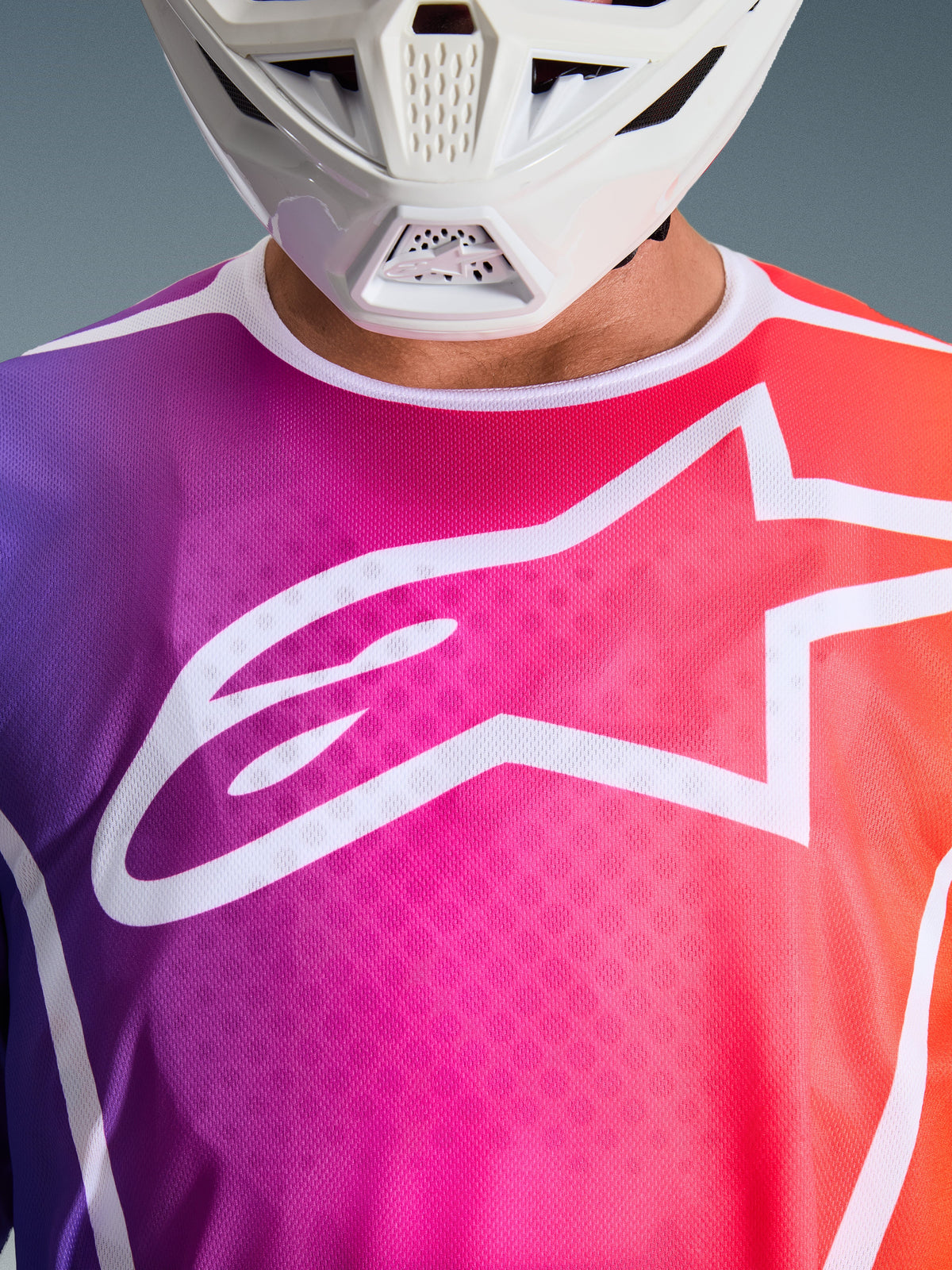 Alpinestars 2026 Fluid Apex Jersey, maillot de motocross, dégradé multicolore violet, rose et orange, avec un grand logo Astars blanc sur la poitrine, vue rapprochée porté avec un casque de motocross blanc