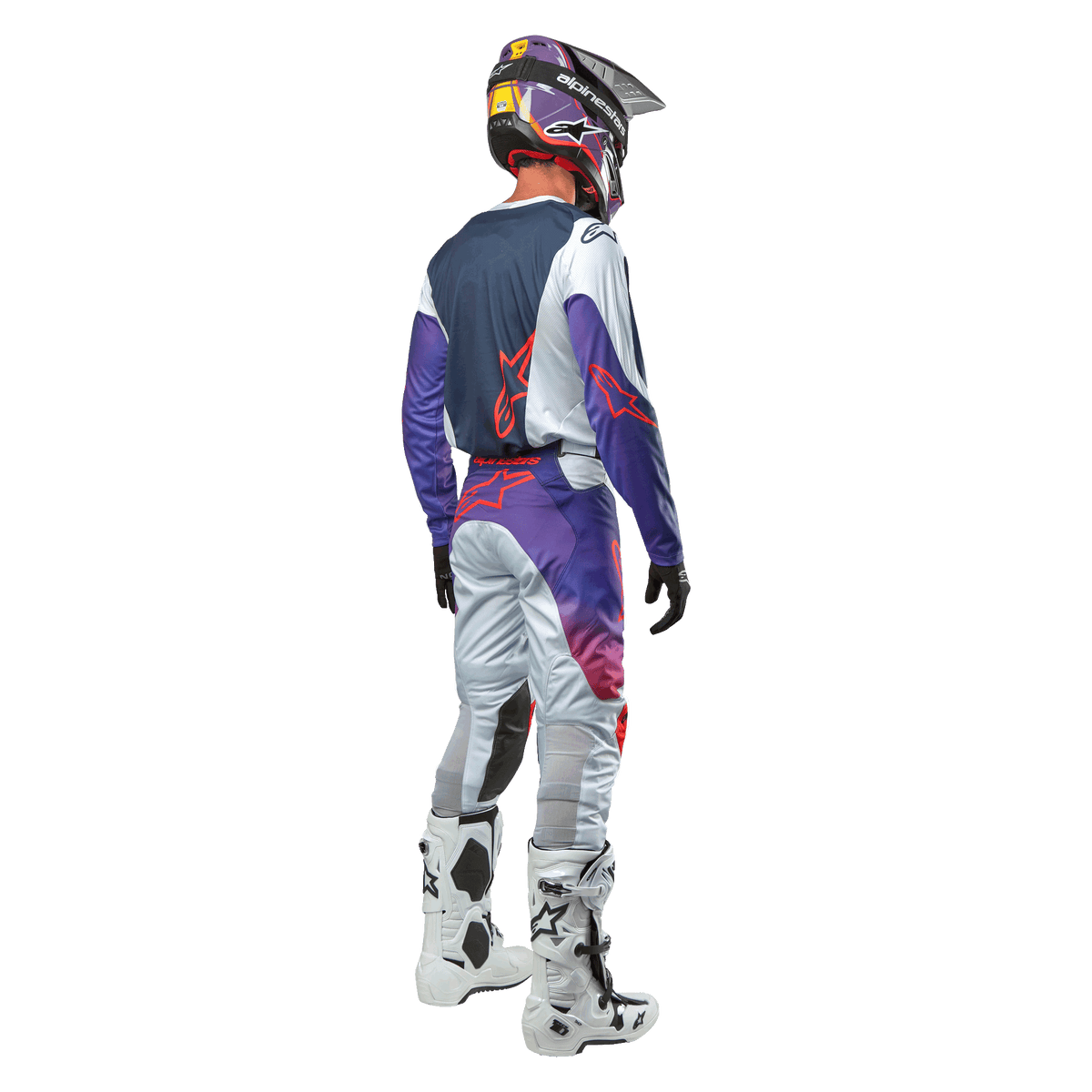 Pilote de motocross vu de dos portant le maillot Alpinestars Racer Hoen 2024 en Light Gray Hot Orange Black, panneau arrière bleu marine avec logo étoile orange, manches violettes, porté avec le pantalon de course assorti et des bottes blanches.
