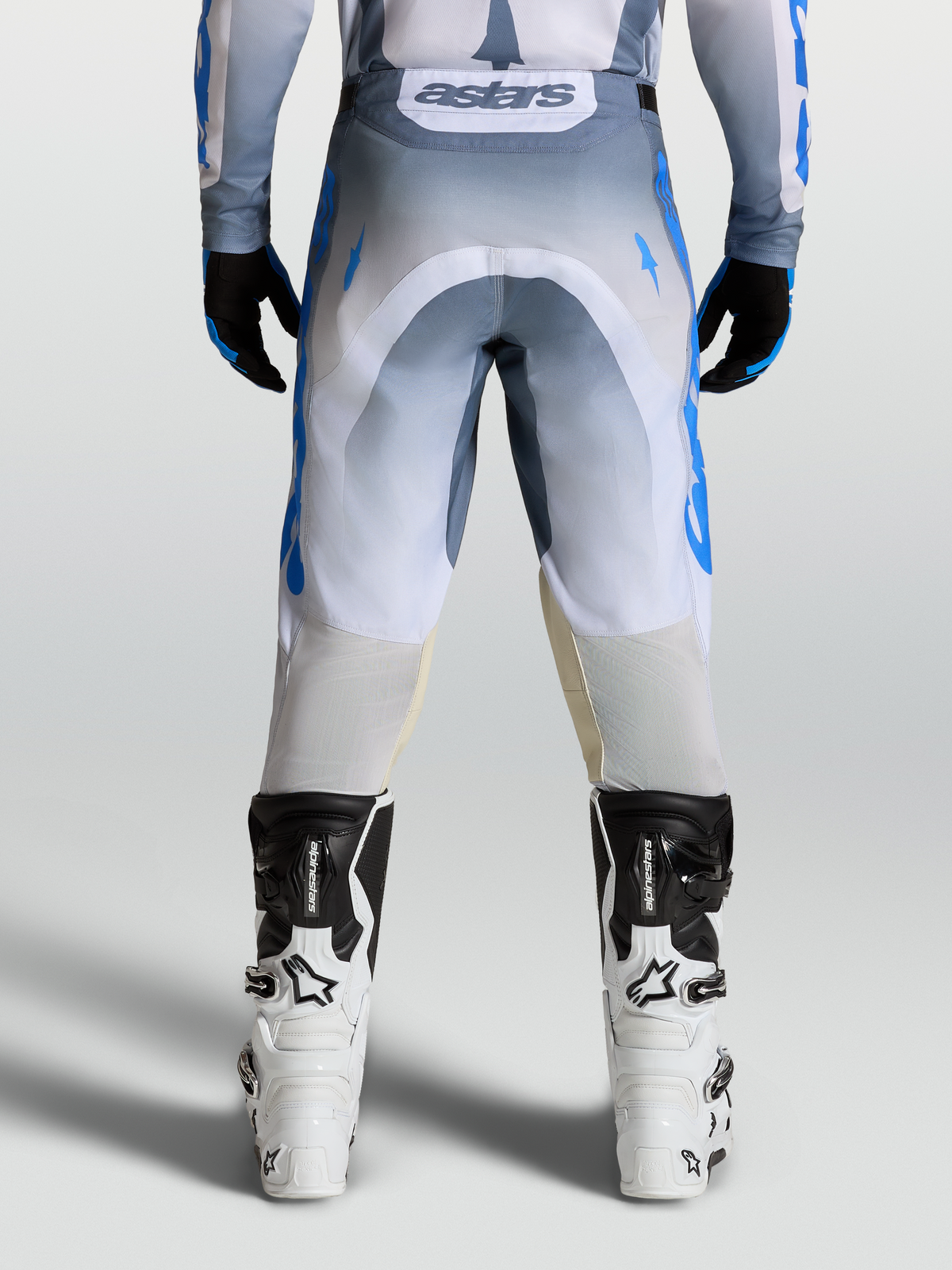 Alpinestars Pantalon Racer Amber, pantalon de motocross pour homme, gris et bleu, vue de dos avec maillot et bottes blanches, construction en poly-tissu 600D durable et inserts extensibles pour le tout-terrain.