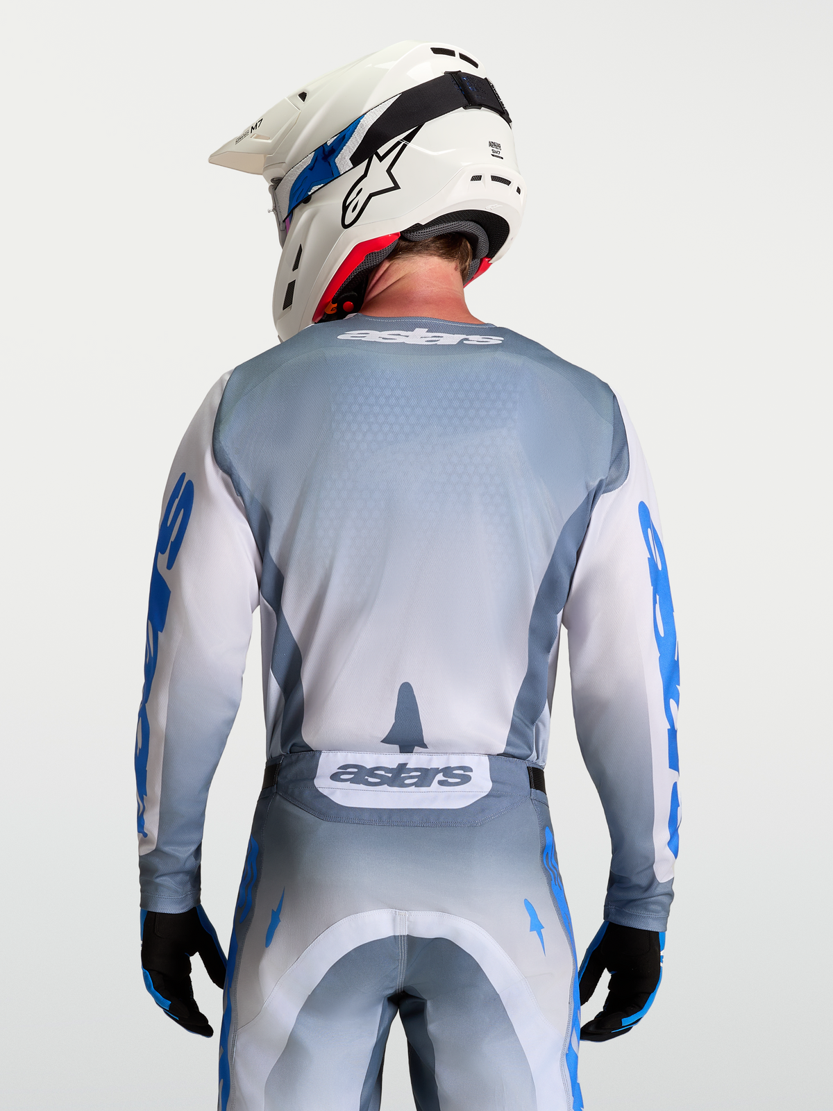 Alpinestars Maillot Racer Amber, maillot de motocross gris et blanc, vue de dos sur mannequin avec casque, graphismes bleu UCLA et lilas, coupe ergonomique racing, tissu poly-matière évacuant l'humidité