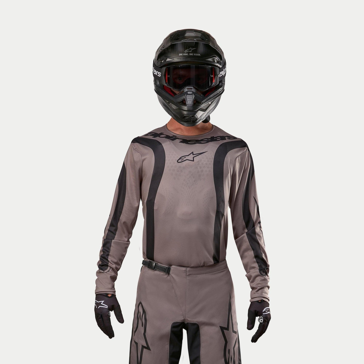 Pilote de motocross portant le maillot Alpinestars Fluid Lurv 2024 en Mud Black, un tissu gris-brun terreux avec des rayures graphiques noires et un logo étoilé sur la poitrine, associé à un casque noir, des lunettes rouges et des gants noirs pour un look de course tout-terrain coordonné.