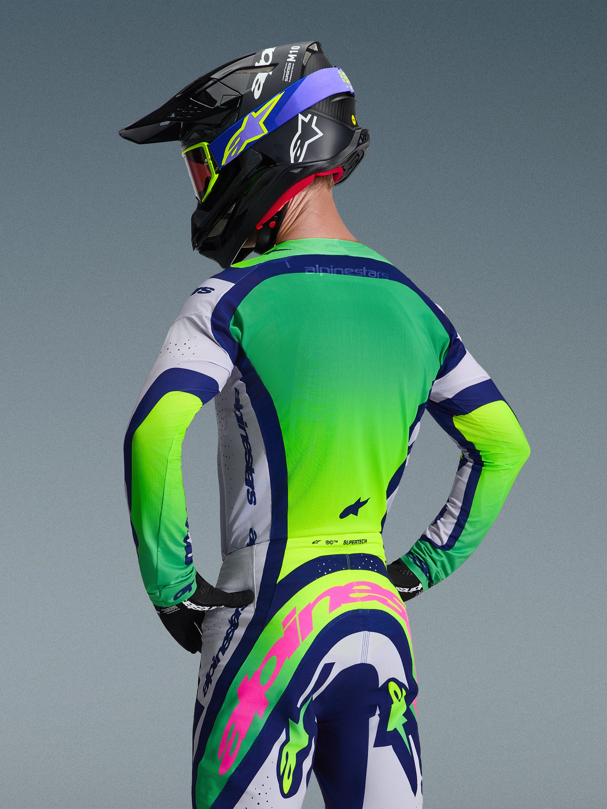 Vue arrière d'un pilote de motocross portant l'ensemble d'équipement Alpinestars Supertech Vista 2026, comprenant un maillot vert et blanc avec des accents bleu marine, un pantalon assorti avec un logo Alpinestars rose proéminent, un casque de course noir avec des graphismes bleus et des lunettes teintées rouges sur fond gris neutre.