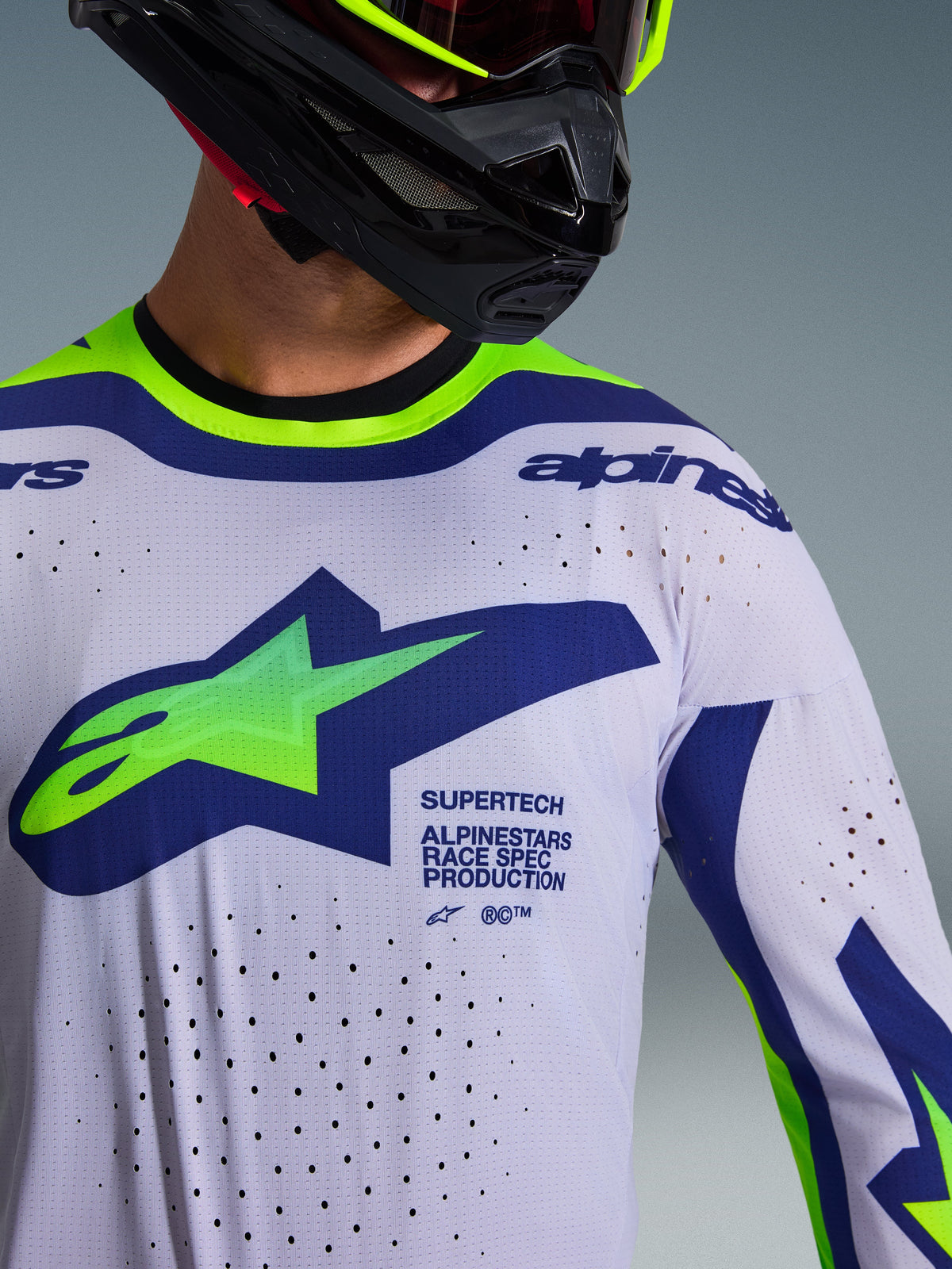 Maillot Alpinestars Supertech Vista 2026 en gris clair, jaune fluo et bleu, gros plan porté par un pilote avec un casque noir, présentant un grand logo sur la poitrine, des perforations de ventilation découpées au laser et un marquage professionnel de course.