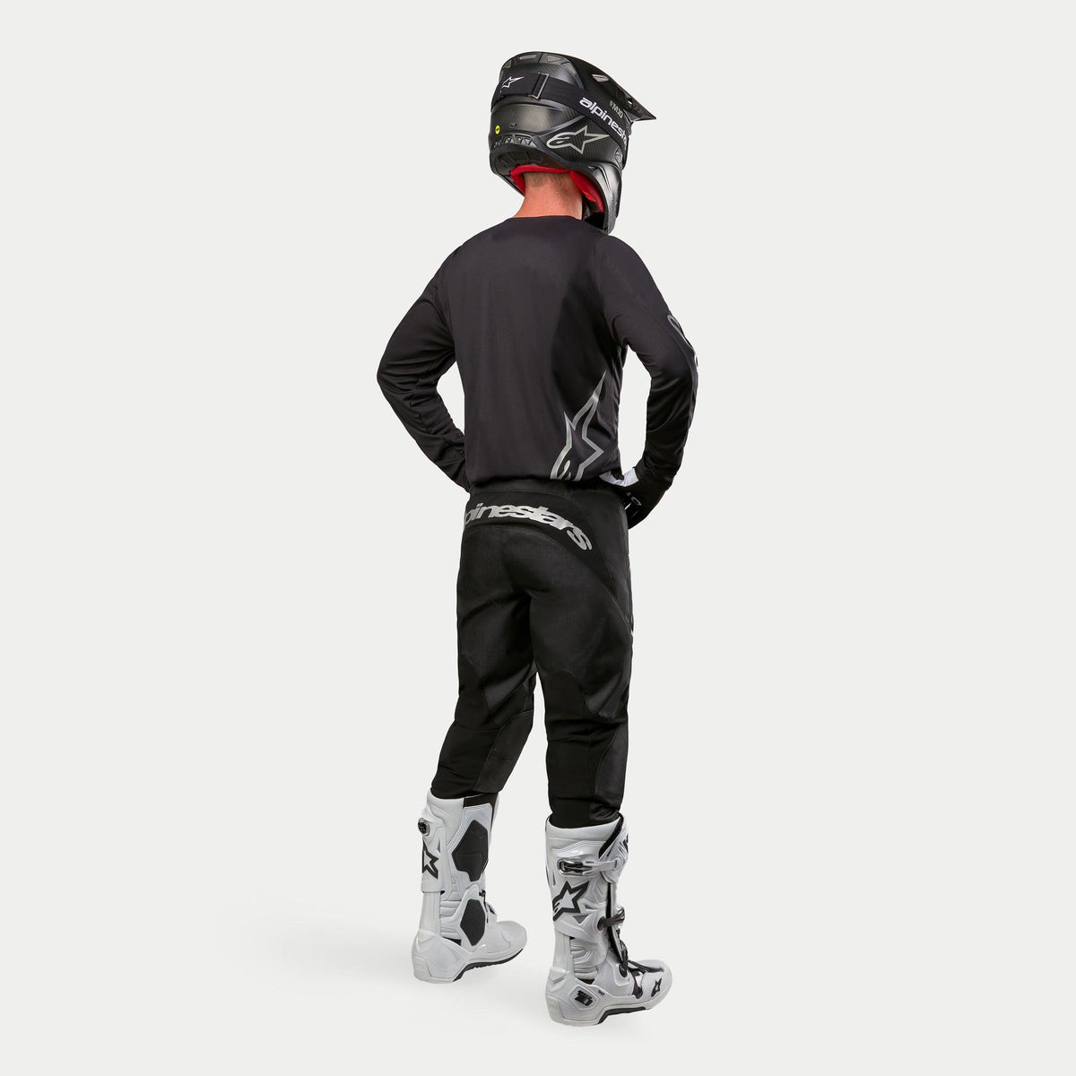 Motocross rider wearing the Alpinestars 2024 Fluid Graphite Jersey in Black Silver from a rear view, long-sleeve athletic fit jersey paired with black motocross pants, black helmet, and white and black boots, complete professional gear for off-road racing.