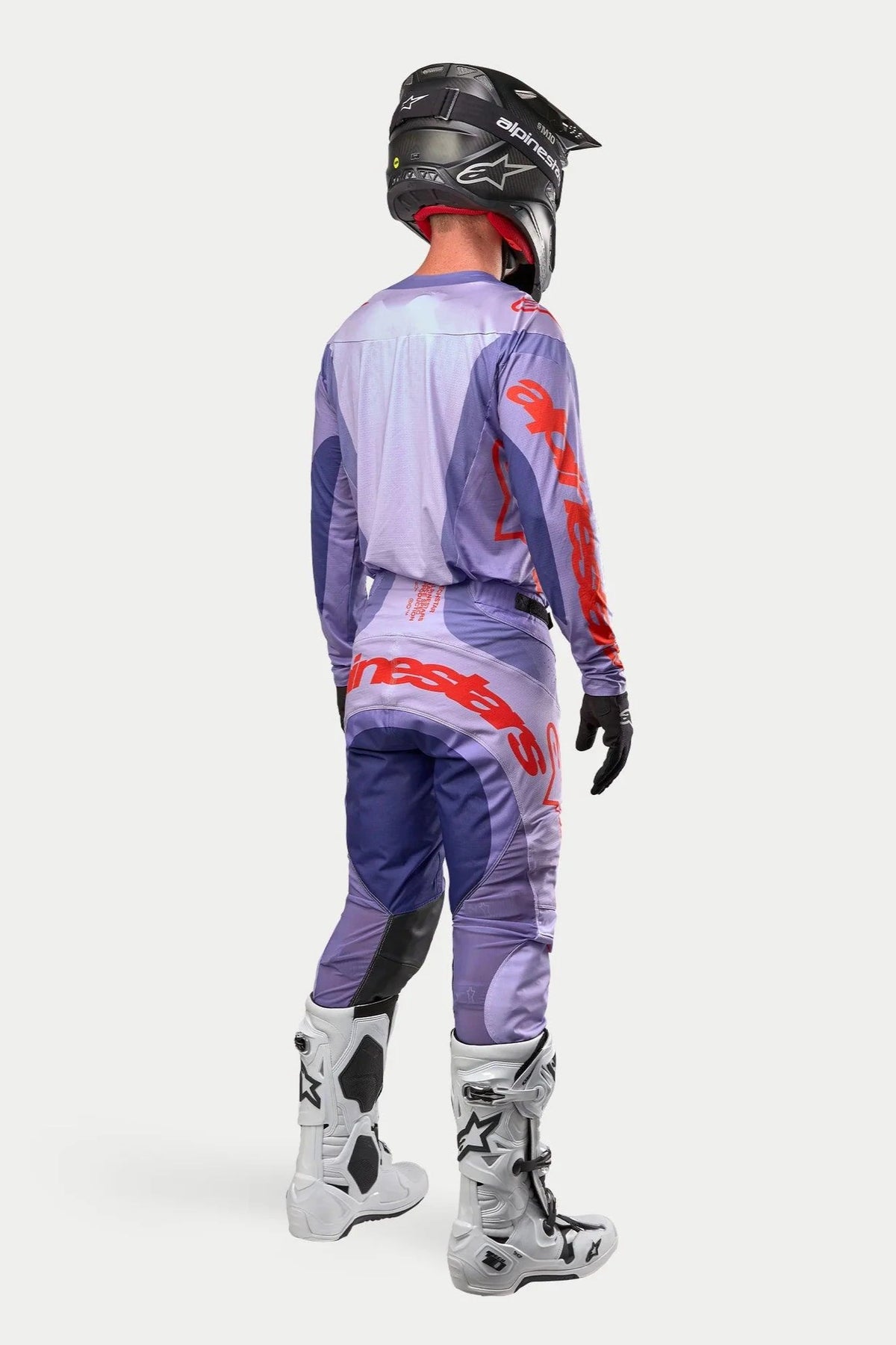 Pilote de motocross portant une tenue de course complète vue de dos, maillot Alpinestars 2024 Techstar Ocuri pourpre avec marquage orange vif, pantalon pourpre assorti avec détails orange, casque noir, bottes blanches et noires, gants noirs, équipement de course tout-terrain professionnel