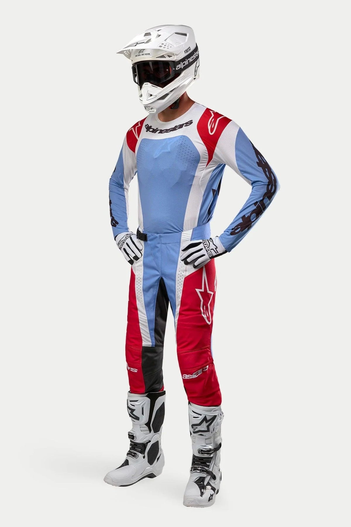 Pilote de motocross portant un équipement de course complet comprenant le maillot Alpinestars 2024 Techstar Ocuri en bleu clair, rouge Mars et blanc, associé à un pantalon assorti, un casque blanc avec des lunettes, des bottes blanches et des gants blancs et noirs.