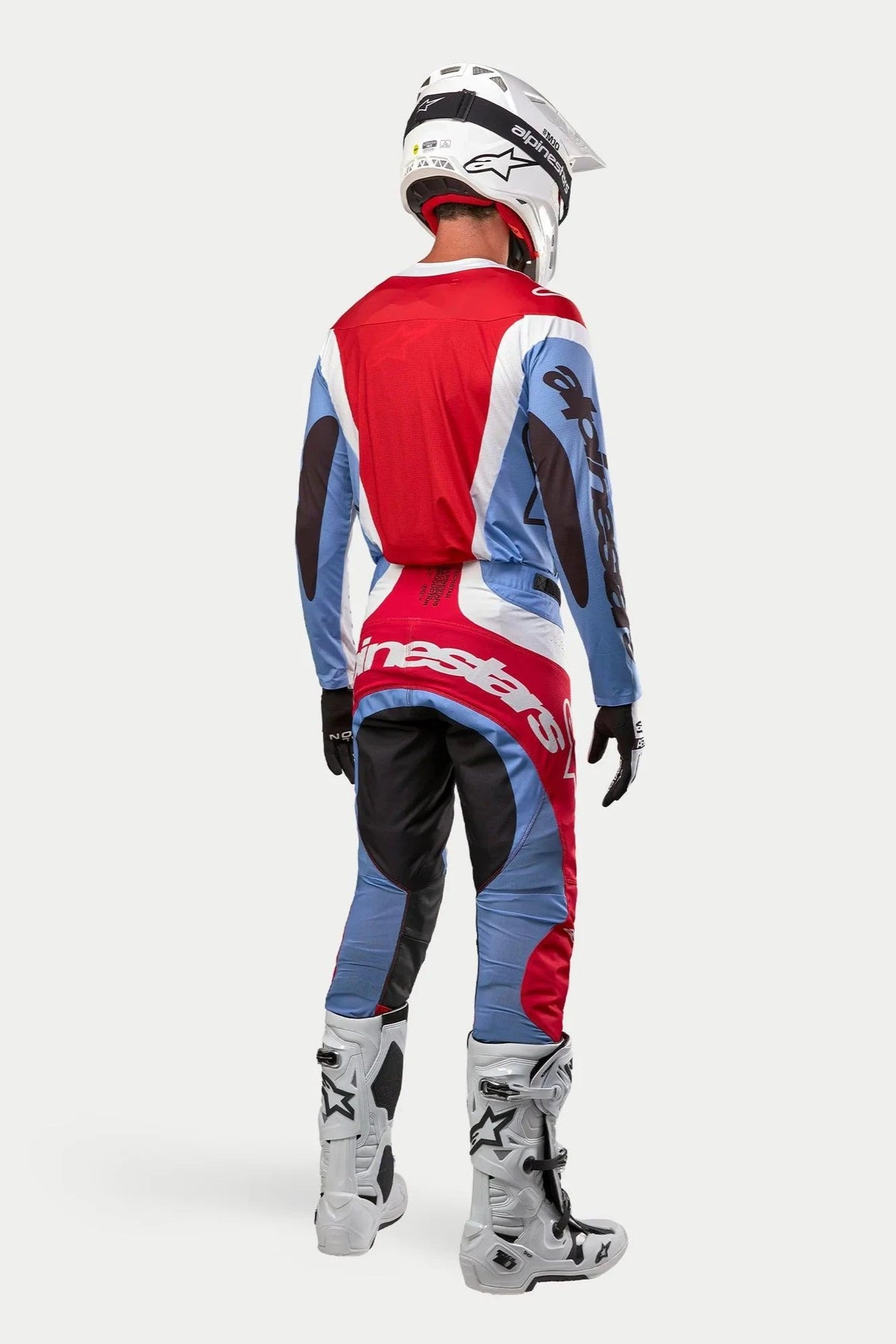 Pilote de motocross de dos portant une tenue de course complète, maillot et pantalon Techstar Ocuri 2024 bleu clair, rouge mars et blanc, casque Alpinestars blanc avec graphismes noirs, bottes tout-terrain blanches et grises, gants noirs, tenue coordonnée pour la course de motocross