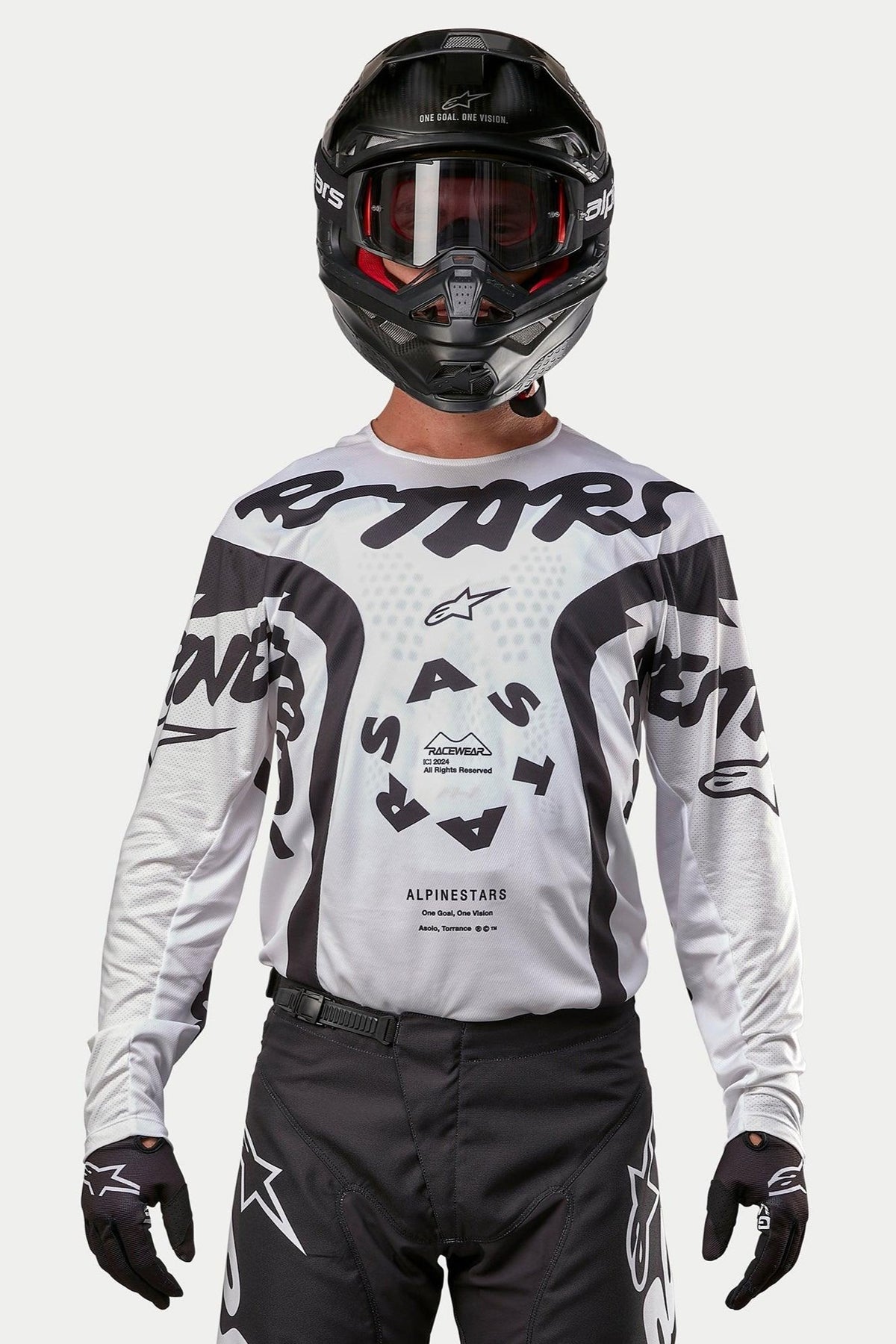 Pilote de motocross portant le maillot Alpinestars 2024 Racer Hana blanc et noir, avec casque et pantalon de motocross noirs, graphismes 'ASTARS' et logo central circulaire, équipement de course tout-terrain professionnel