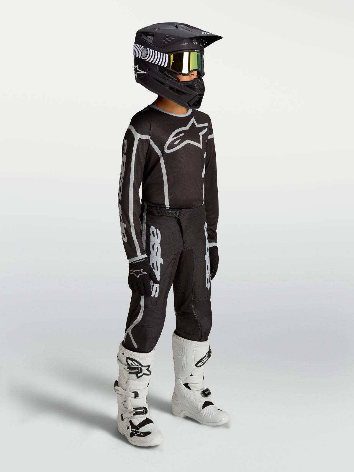 Alpinestars maillot Fluid Apex V2 Junior et ensemble de motocross pour enfant, noirs et argentés, textile respirant, porté par un jeune pilote avec des bottes blanches.