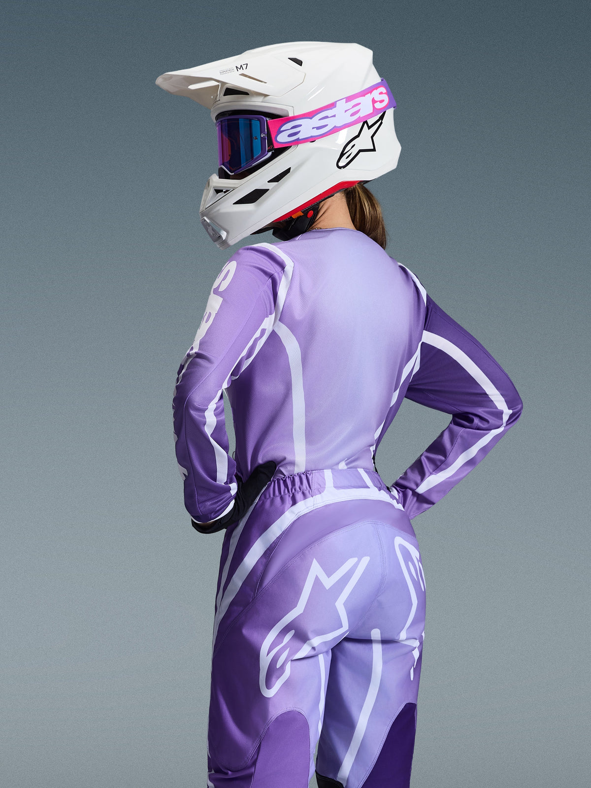 Motocross rider wearing Alpinestars 2026 Stella Fluid Apex Gear Set, Purple and lavender jersey and pants with white graphics, white helmet with pink and purple goggles, dedicated women's fit, coordinated gear for off-road racing