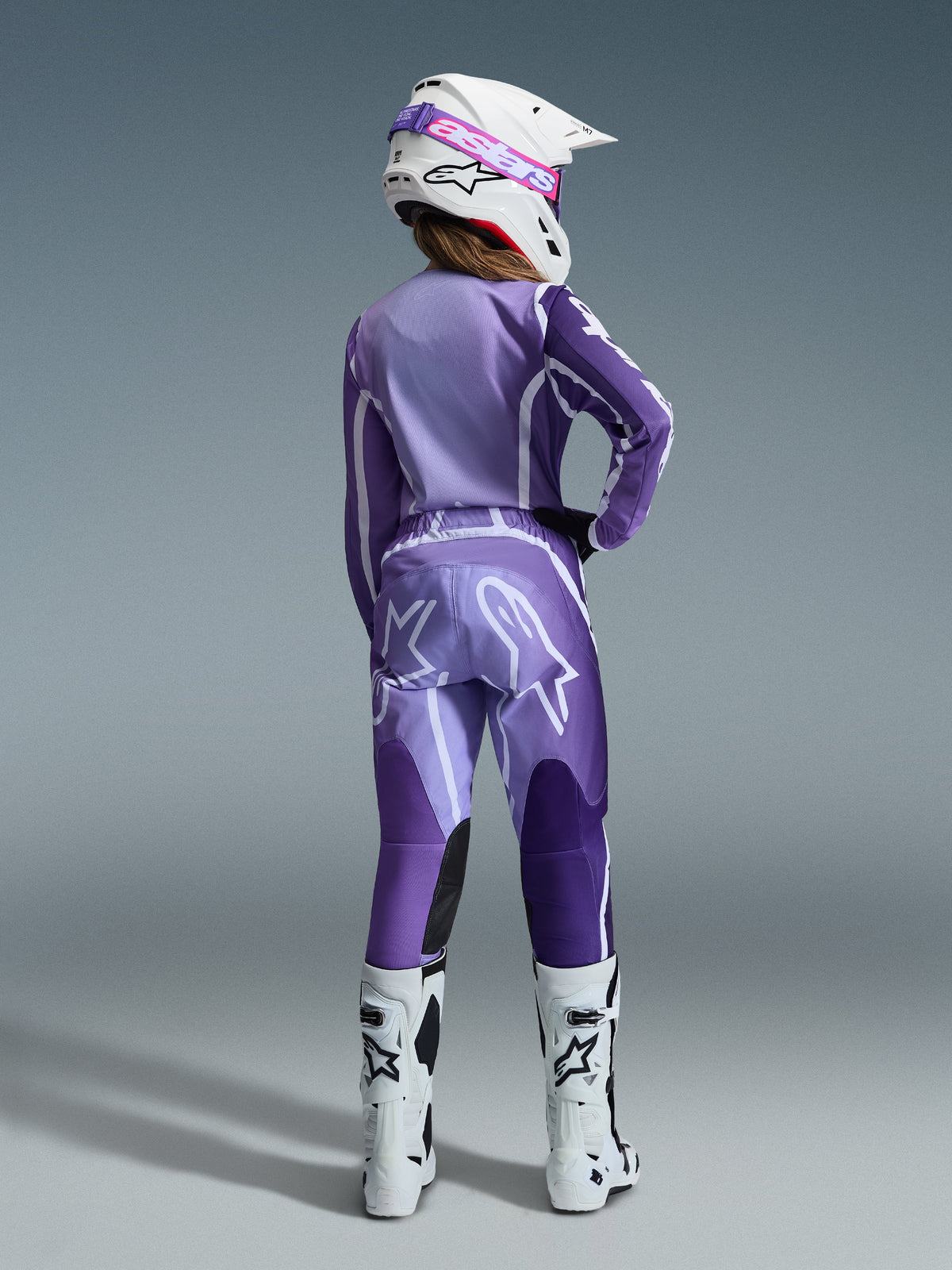 Alpinestars 2026 Stella Fluid Apex Gear Set, Women's Motocross Gear, Purple and Lavender with white accents, female rider seen from the back wearing matching jersey and pants, white helmet with pink goggles, and white boots, coordinated athletic racing outfit