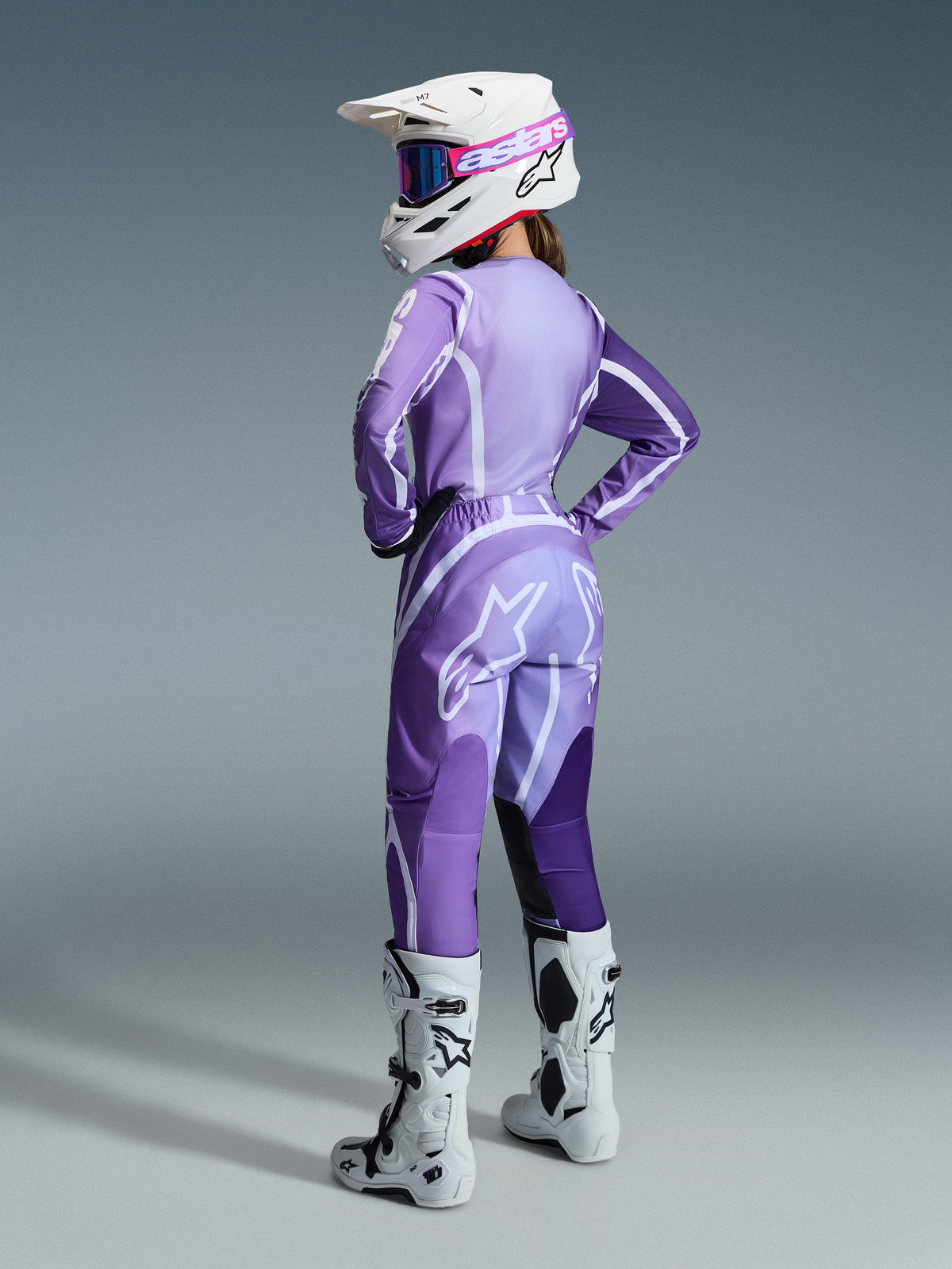 Female motocross rider wearing the 2026 Woman Stella Fluid Apex Gear Set, purple and lavender jersey and pants with white accents, white helmet with pink goggles, white and black boots, coordinated outfit for off-road racing