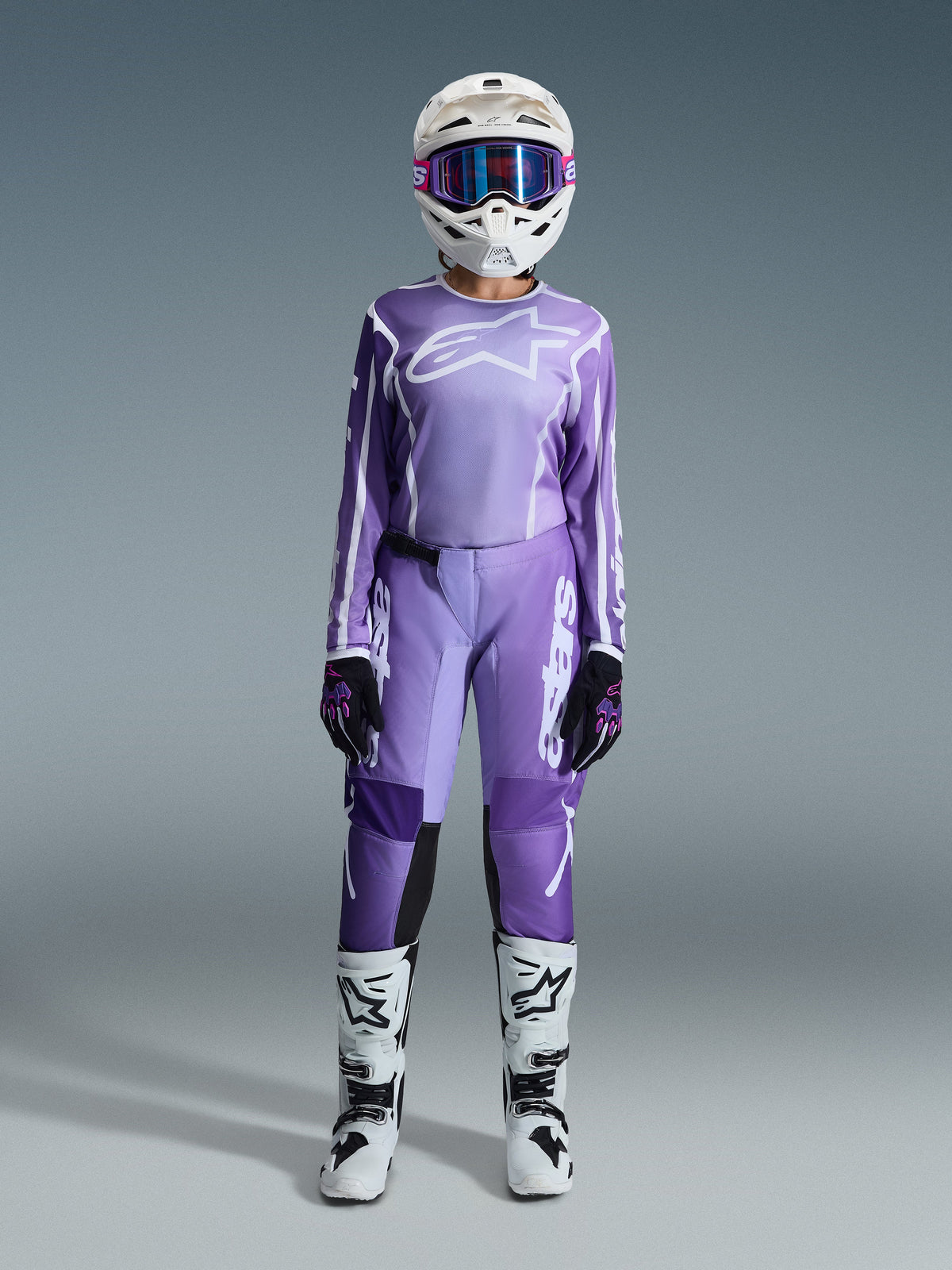 Motocross rider wearing the Alpinestars 2026 Stella Fluid Apex Gear Set, purple jersey with white logo, matching purple pants with white branding, white helmet with pink goggles, white and black boots, coordinated women's motocross outfit for off-road racing