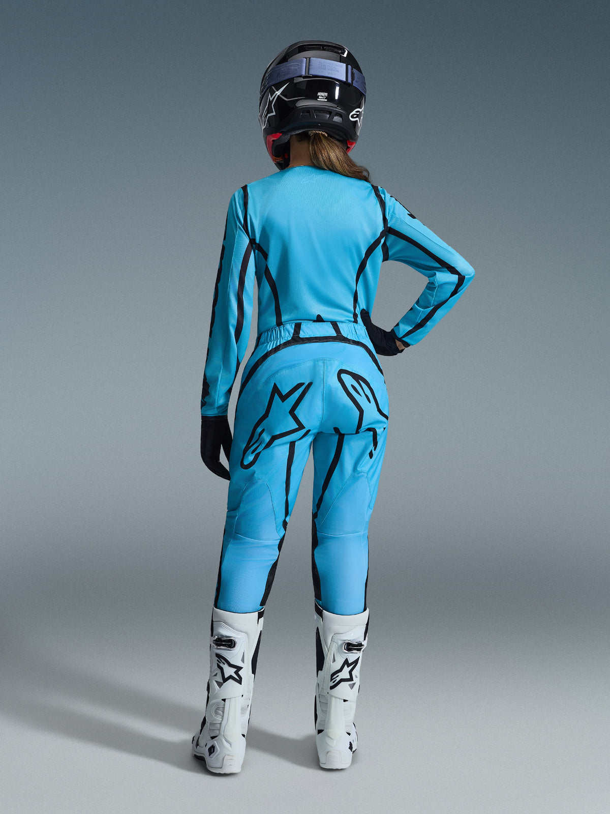 Pilote de motocross femme vue de dos, portant l'ensemble Alpinestars Stella Fluid Apex 2026 pour femme, bleu vif avec des accents de lignes graphiques noires, grand logo étoile noir sur le pantalon, bottes blanches et noires et casque noir.