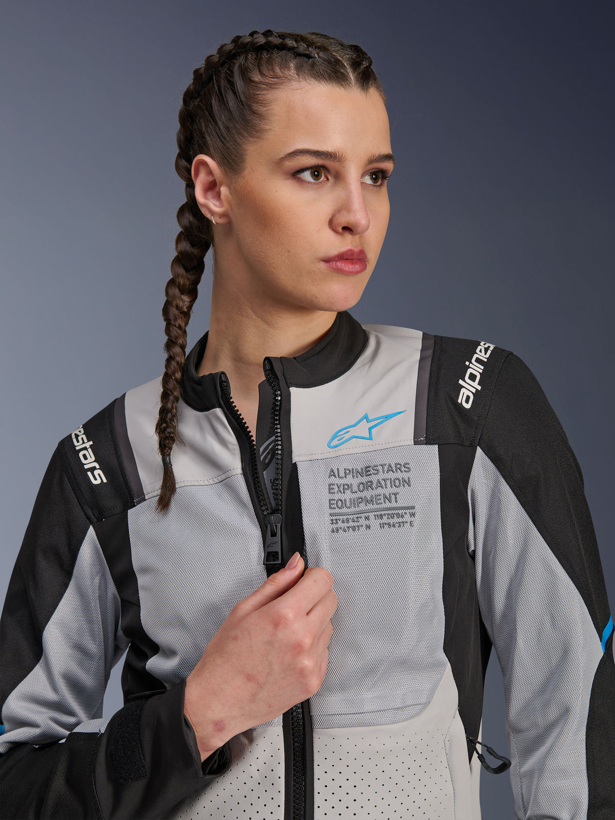 Femme portant la veste Alpinestars Stella ST-2 Air en Ice Gray et noir, portrait en gros plan, avec des panneaux en mesh pour la ventilation, marquage « ALPINESTARS EXPLORATION EQUIPMENT » sur la poitrine avec un logo bleu, veste de moto d'aventure moderne