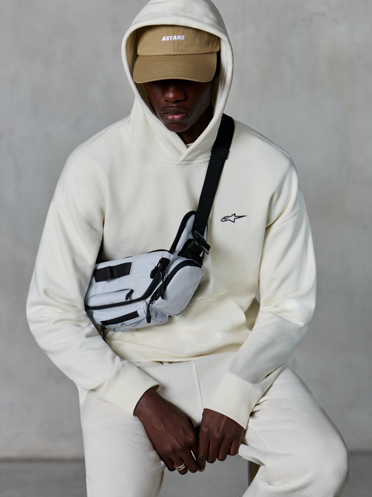 Sweat à capuche Alpinestars Essentials Ageless, streetwear décontracté, blanc cassé, petit logo noir brodé sur la poitrine, capuche portée sur une casquette beige avec marquage ASTARS, stylisé avec un sac bandoulière gris clair, vêtement lifestyle premium