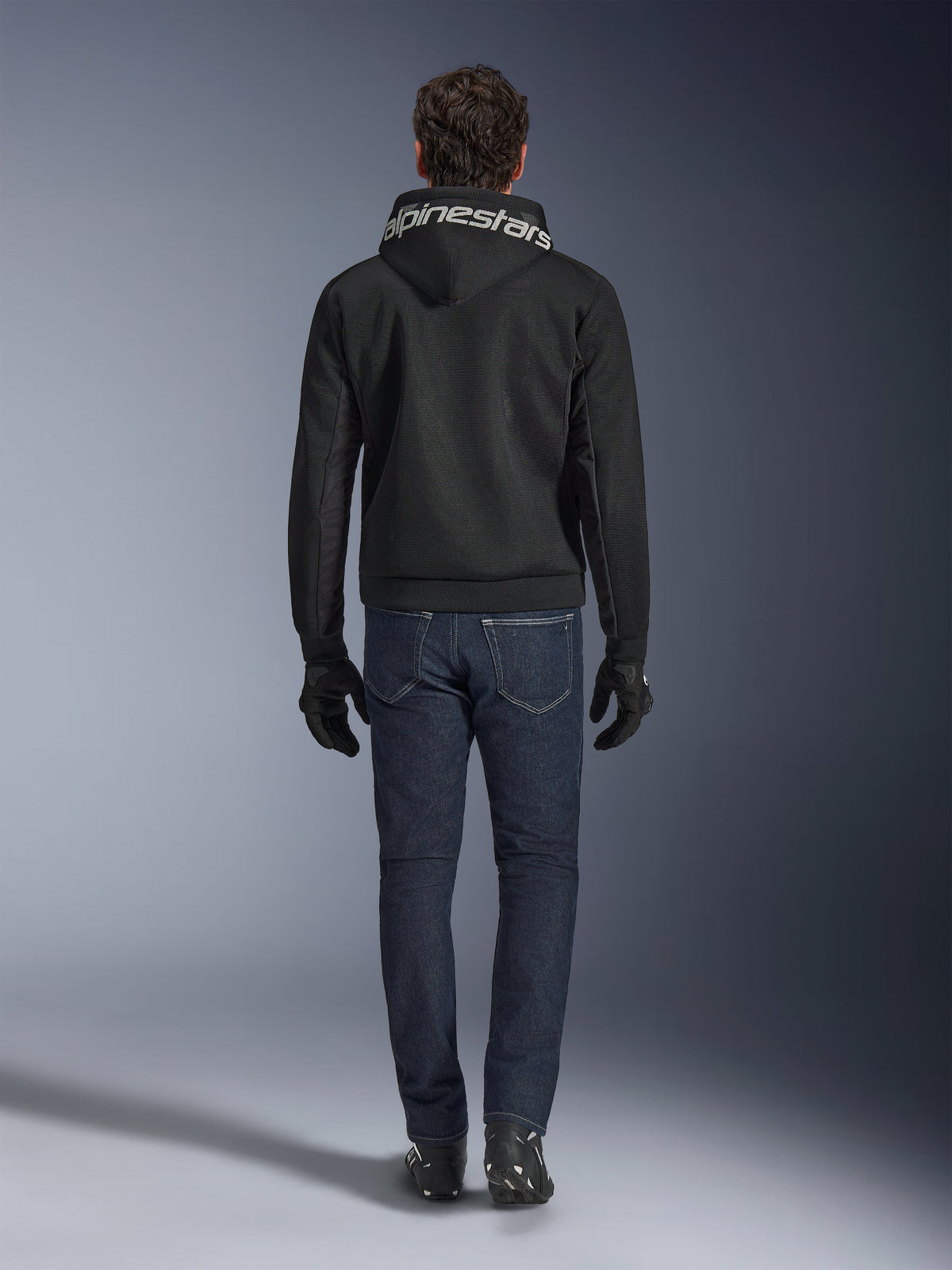 Alpinestars Chrome Superairflow Hoodie, sweat à capuche de moto noir avec logo blanc sur la capuche, vue de dos, porté avec un jean bleu foncé, des gants noirs et des bottes noires et blanches, équipement de conduite sportive urbaine