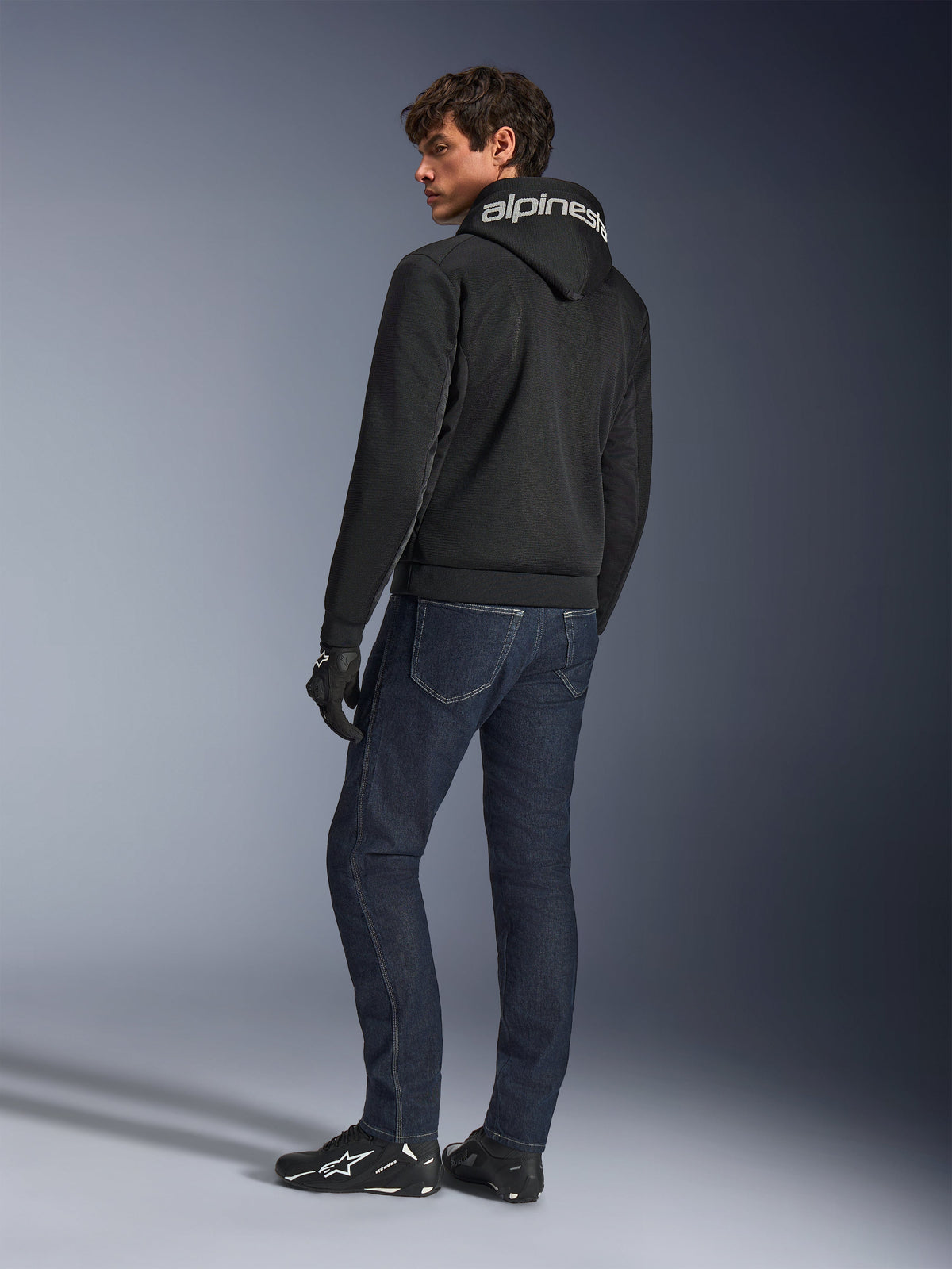 Hoodie Alpinestars Chrome Superairflow, noir, vue de dos sur mannequin homme, avec logo Alpinestars blanc sur la capuche, porté avec un jean en denim foncé et des gants noirs, design d'équipement de moto urbain