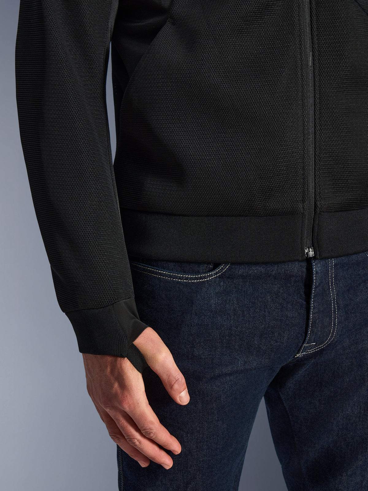 Alpinestars Chrome Superairflow Hoodie, noir, gros plan sur le tissu technique en maille 3D haute résistance, avec poignet en jersey côtelé extensible et boucle de rétention élastique pour le pouce, porté avec un jean bleu