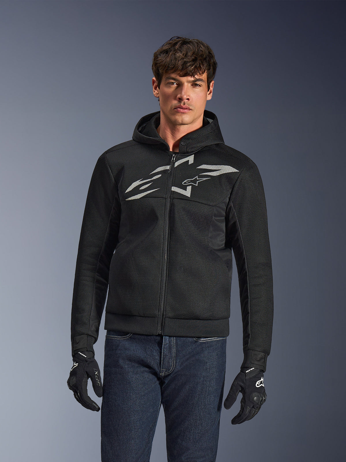 Alpinestars Chrome Superairflow Hoodie, sweat à capuche de moto noir à fermeture éclair avec grand logo blanc sur la poitrine, construction en maille aérée, porté avec un jean bleu et des gants noirs, vêtement de conduite urbaine sportive
