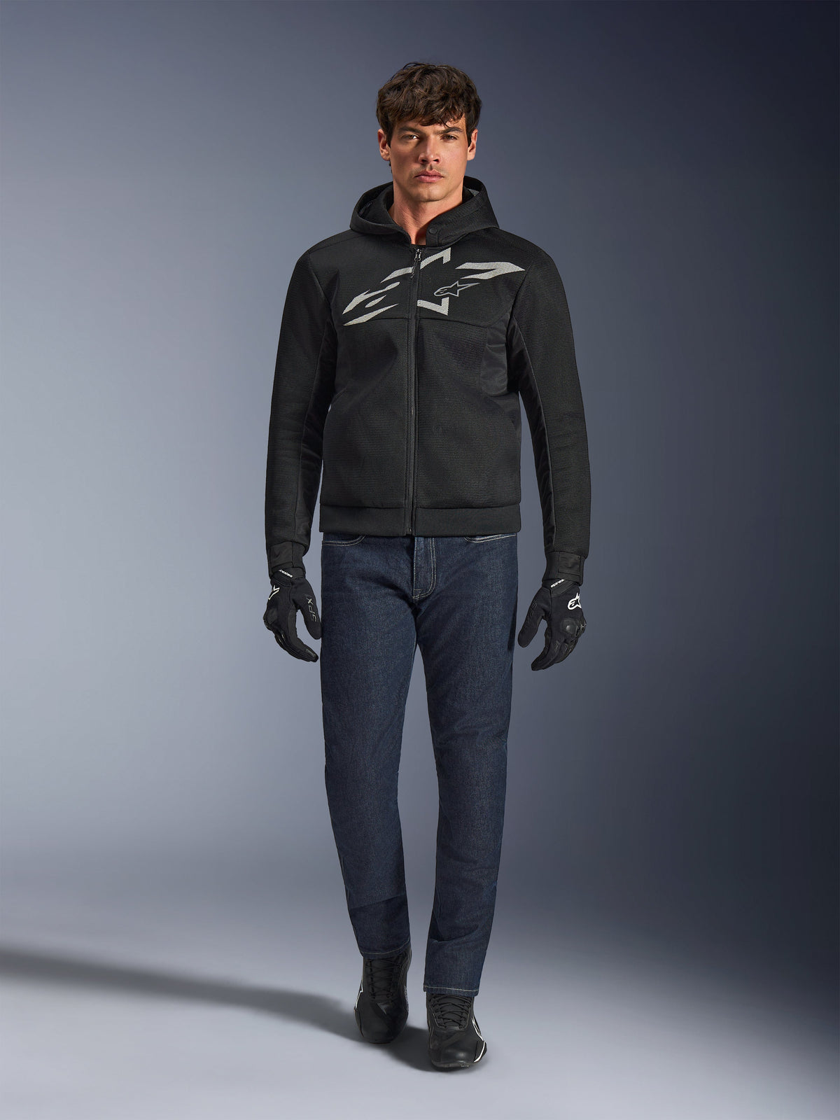 Motard portant le hoodie Alpinestars Chrome Superairflow noir et gris, associé à un jean bleu foncé, des gants de moto noirs et des chaussures noires. Le sweat à capuche présente un grand logo stylisé sur la poitrine et une texture en maille 3D pour une ventilation optimale par forte chaleur.