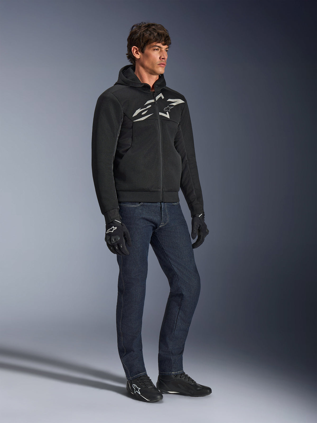 Hoodie Alpinestars Chrome Superairflow, noir avec logo blanc et gris, construction en textile maille 3D ventilée, porté avec un jean en denim bleu foncé et des gants de moto noirs, style de conduite sport urbain