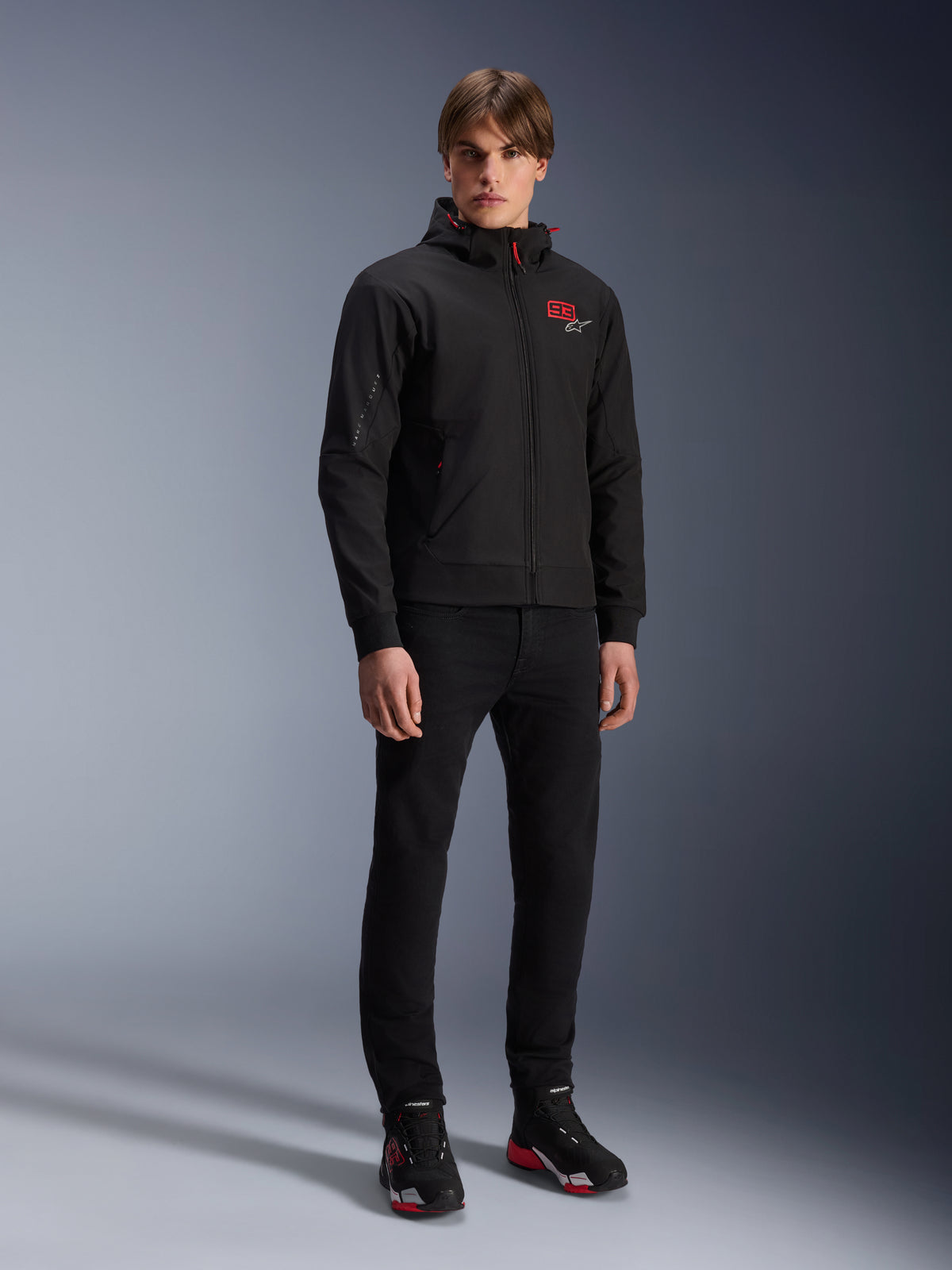 Alpinestars MM93 Radium Tech Hoodie, Urban Motorcycle Jacket, Black with Bright Red accents, worn by model with black pants and riding shoes, featuring red '93' chest logo and Marc Marquez branding on the sleeve, casual technical streetwear for urban riding