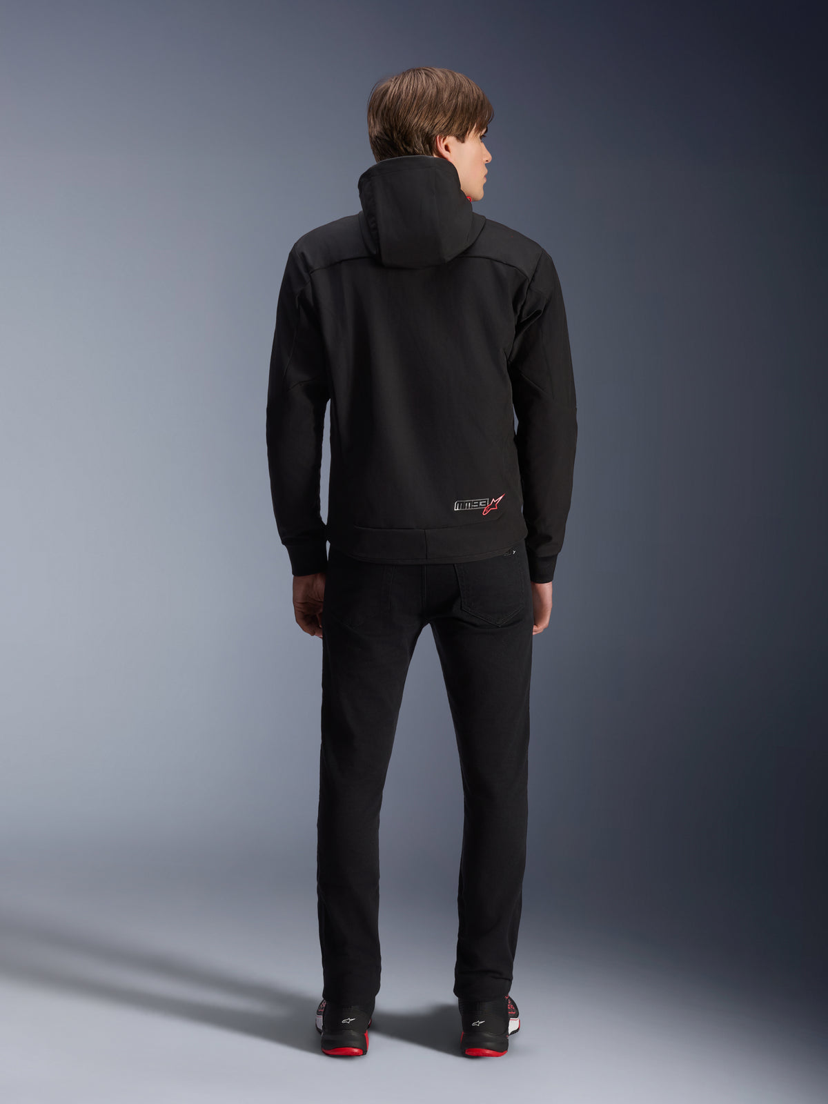 Alpinestars MM93 Radium Tech Hoodie, Black, rear view on model, featuring a small MM93 logo with red star on the lower back, paired with black pants, urban motorcycle riding apparel
