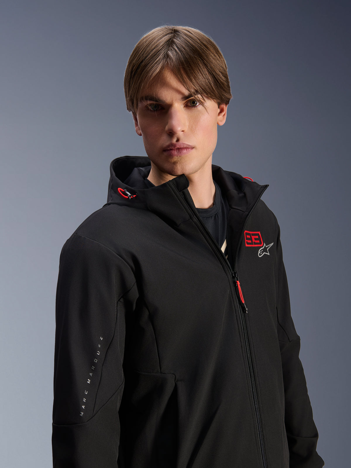 Male model wearing Alpinestars MM93 Radium Tech Hoodie, black urban motorcycle jacket, featuring red '93' and white star logo on the chest, 'Marc Marquez' branding on the sleeve, technical windproof shell for urban riding.