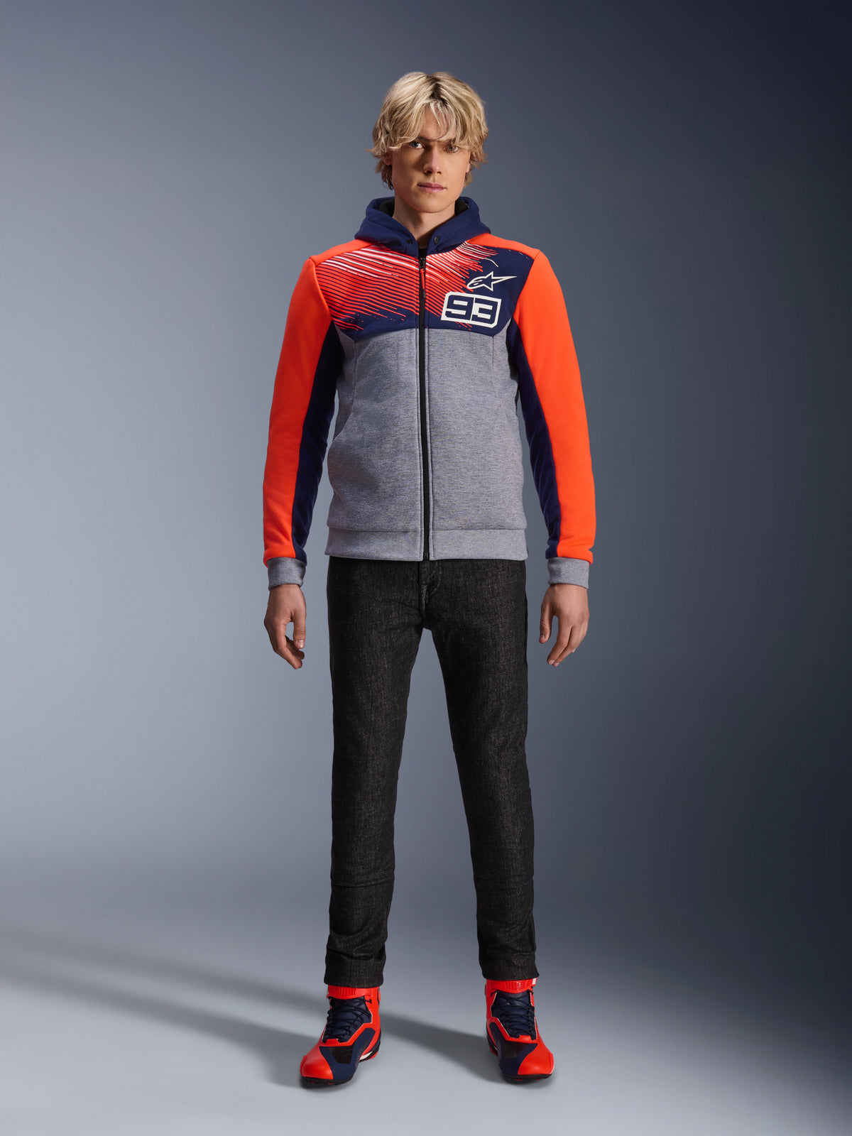 Sweat à capuche Alpinestars MM93 Chrome V2 Sport, sweat à capuche de protection pour la moto, bleu marine, gris chiné et rouge fluo, porté avec un pantalon de conduite sombre et des baskets aux accents rouges, doté d'une fermeture éclair sur toute la longueur et du logo MM93, design urbain décontracté inspiré du sport automobile