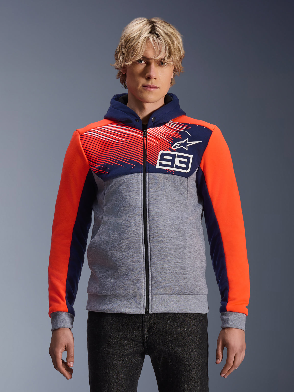 Alpinestars MM93 Chrome V2 Sport Hoodie, sweat à capuche moto zippé, gris chiné avec poitrine et capuche bleu marine, manches orange vif, design numéro 93 sur la poitrine avec motif rayé et logo étoile, porté par un mannequin avec un jean foncé, collection Marc Marquez.