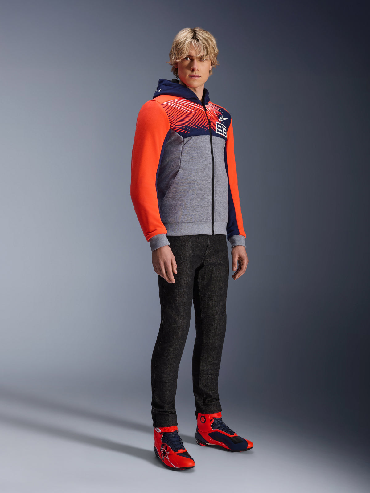 Sweat à capuche sport Alpinestars MM93 Chrome V2, sweat de moto urbain, corps gris chiné avec poitrine bleu marine et manches rouge fluo, logo 93 sur la poitrine, porté par un mannequin avec un pantalon noir ajusté et des chaussures de moto montantes coordonnées rouges et bleues, coupe athlétique moderne pour la conduite urbaine
