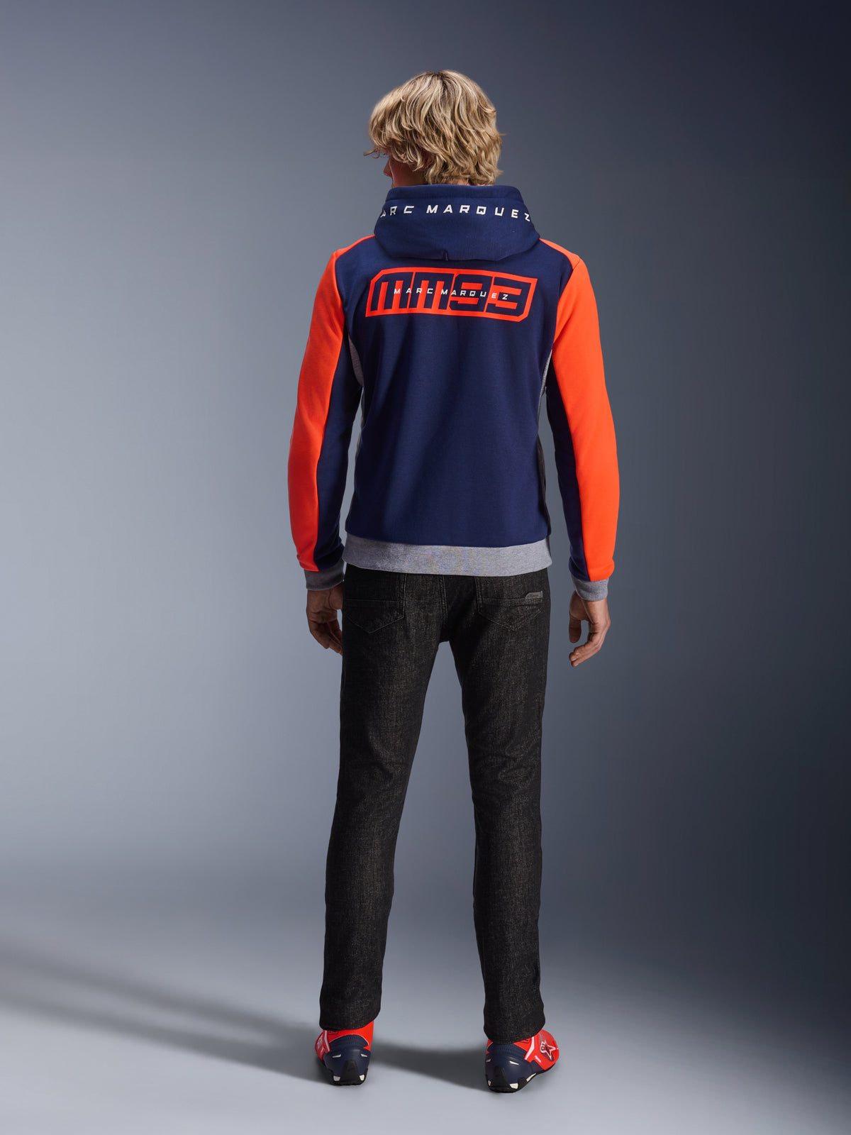 Modèle de dos portant le sweat à capuche Alpinestars MM93 Chrome V2 Sport, bleu marine avec manches rouge fluo éclatant et détails gris chiné, grands logos MM93 et Marc Marquez dans le dos et sur la capuche, sweat à capuche de moto décontracté associé à un pantalon noir