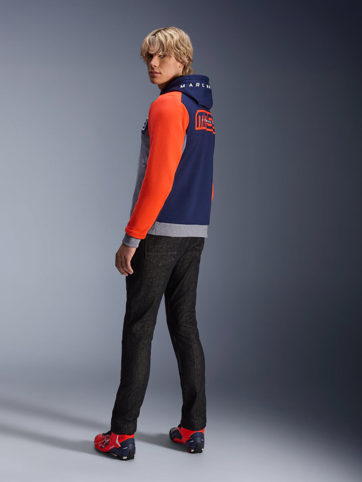 Alpinestars MM93 Chrome V2 Sport Hoodie, bleu marine, gris chiné et rouge fluo, mannequin portant le sweat à capuche avec des manches rouge vif et le logo MM93 Marc Marquez au dos, associé à un jean foncé et des chaussures de moto rouges, équipement de moto urbain décontracté