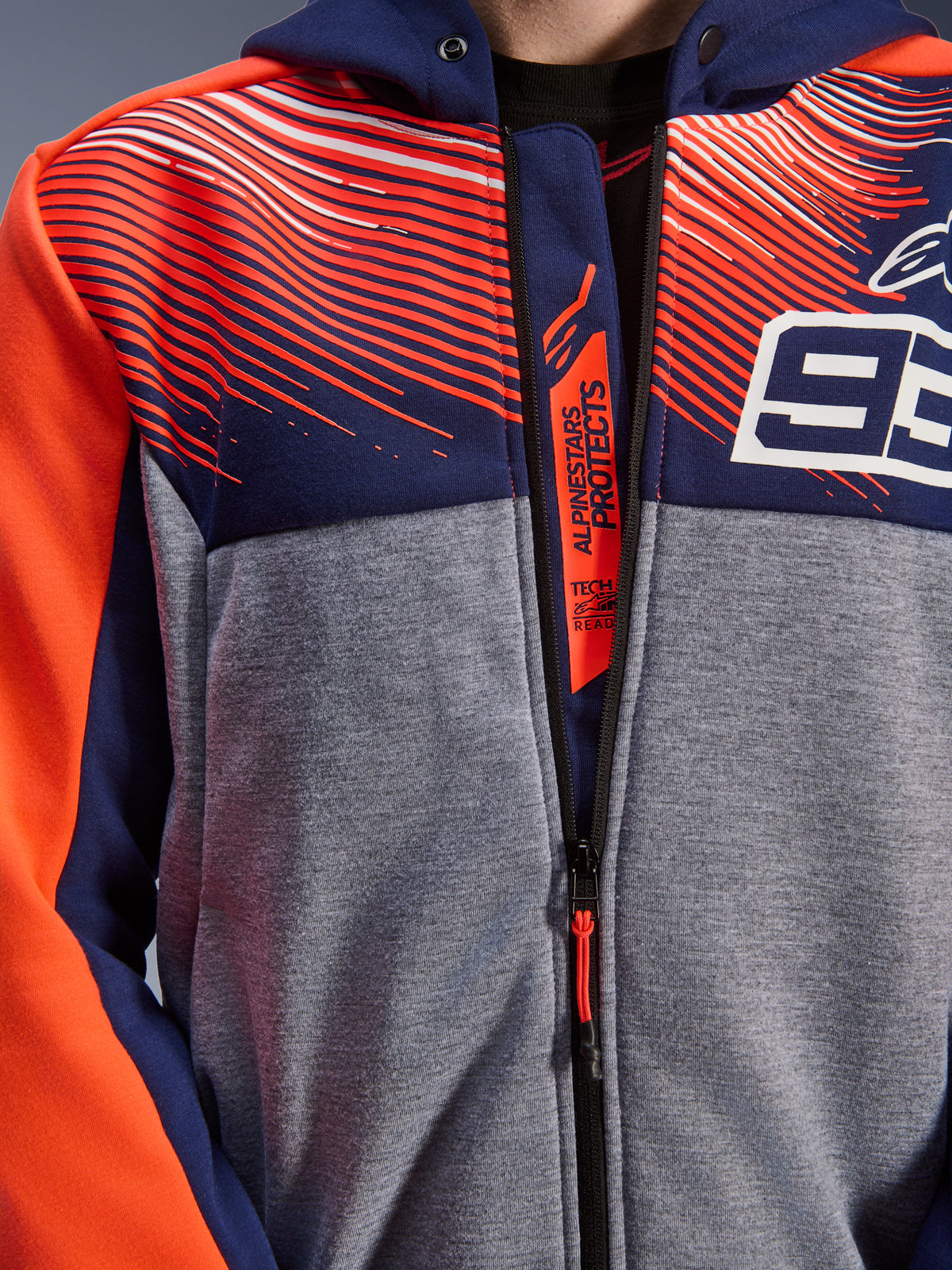 Alpinestars MM93 Chrome V2 Sport Hoodie, gros plan de la poitrine et du col, polaire technique bleu marine et gris chiné avec graphismes rouge fluo, avec le logo 93 blanc et le marquage Alpinestars Protects sur le rabat intérieur de la fermeture éclair, vêtement de moto Tech-Air Ready.