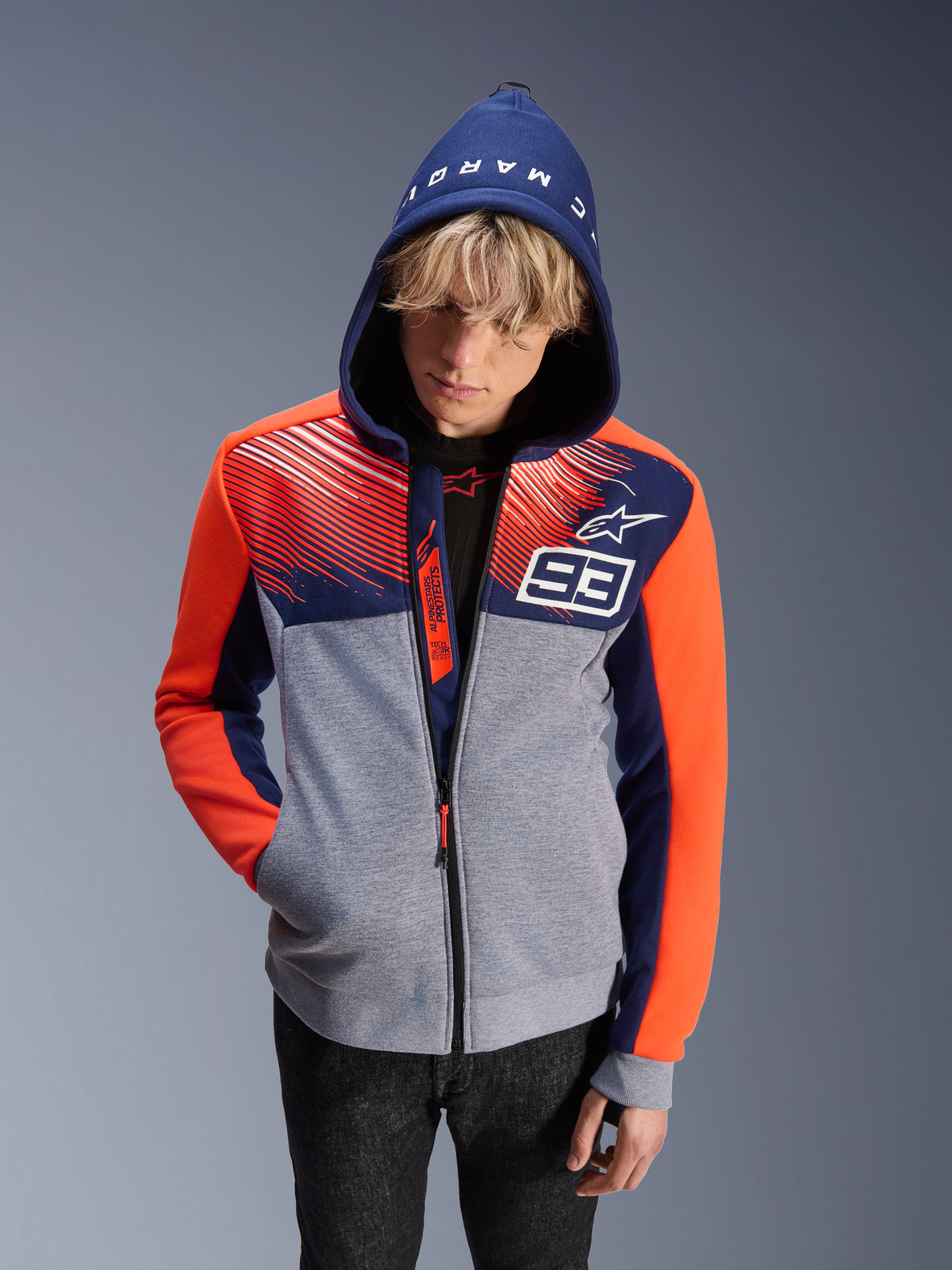 Alpinestars MM93 Chrome V2 Sport Hoodie, sweat à capuche de moto, corps gris chiné avec haut bleu marine et manches orange fluo, numéro 93 blanc et logo Alpinestars, texte Marc Marquez sur la capuche, porté par un modèle masculin avec capuche relevée, design urbain inspiré des sports mécaniques