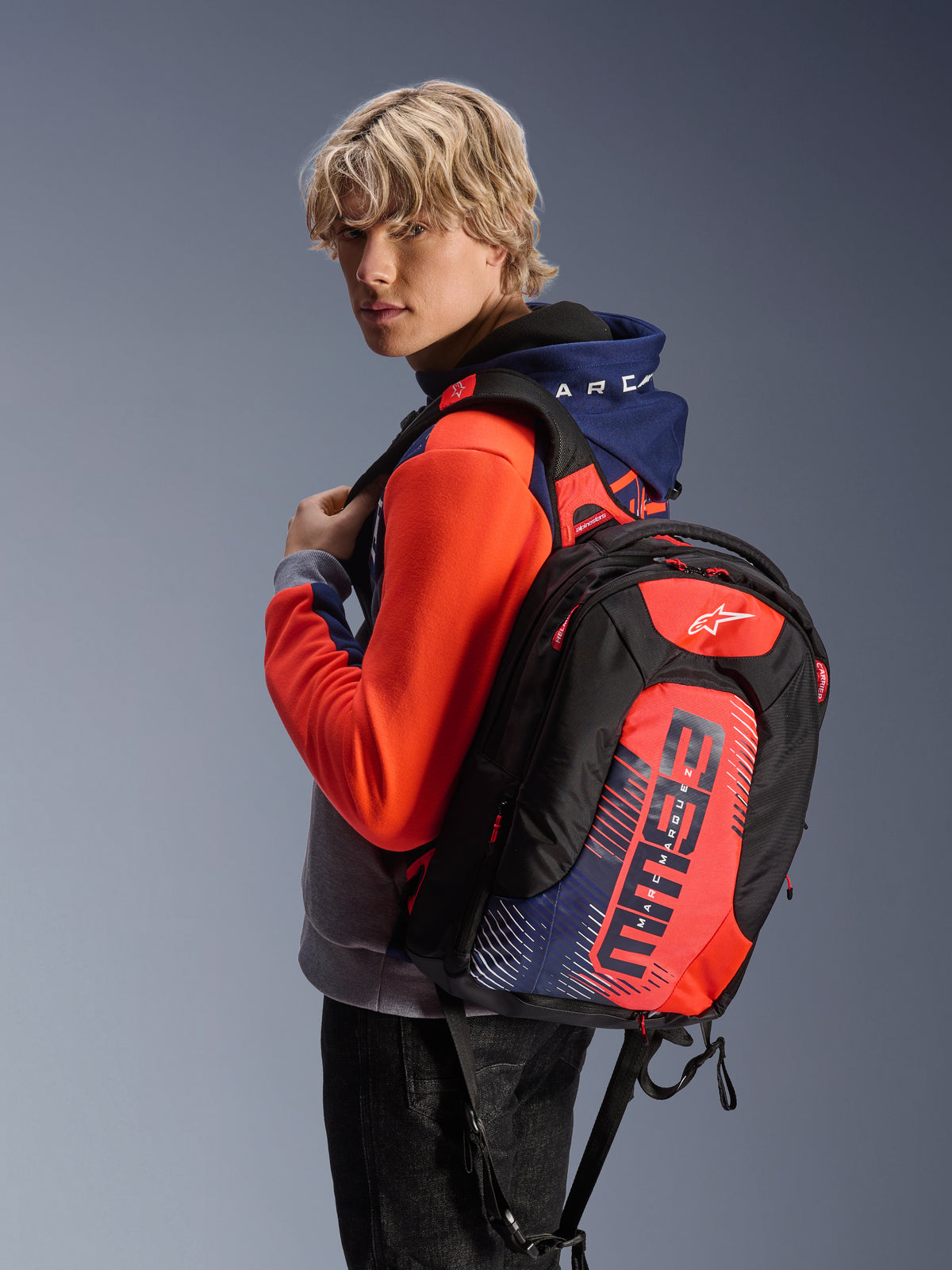 Alpinestars MM93 City Hunter V2 Backpack, black with red fluo and navy blue accents, featuring Marc Marquez MM93 signature graphics, worn with a coordinating sport hoodie, sleek aerodynamic profile for motorcycle commuting