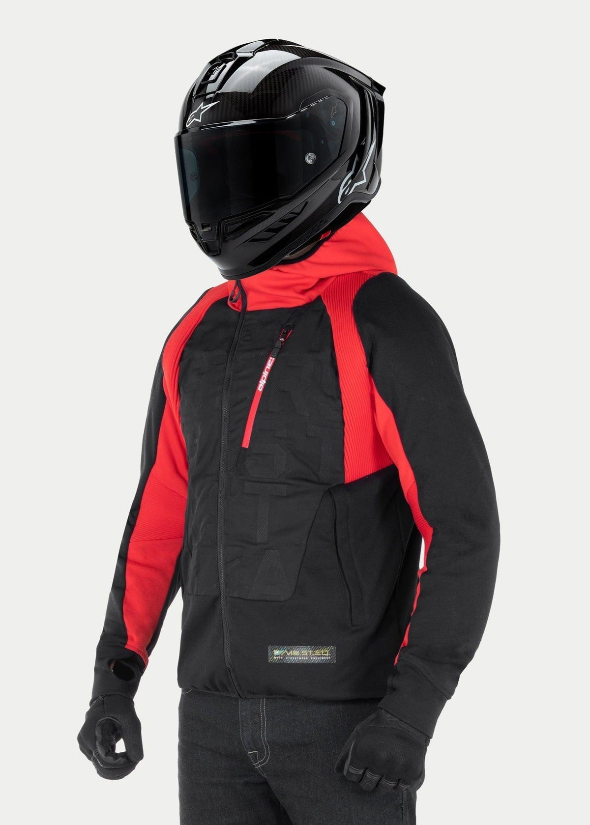 Alpinestars MO.ST.EQ Hybrid Hooded Jacket, sweat à capuche de moto urbain, noir et rouge, avec fermeture éclair avant inclinée et empiècements côtelés aux épaules, portée avec un casque intégral noir et des gants noirs, style streetwear contemporain