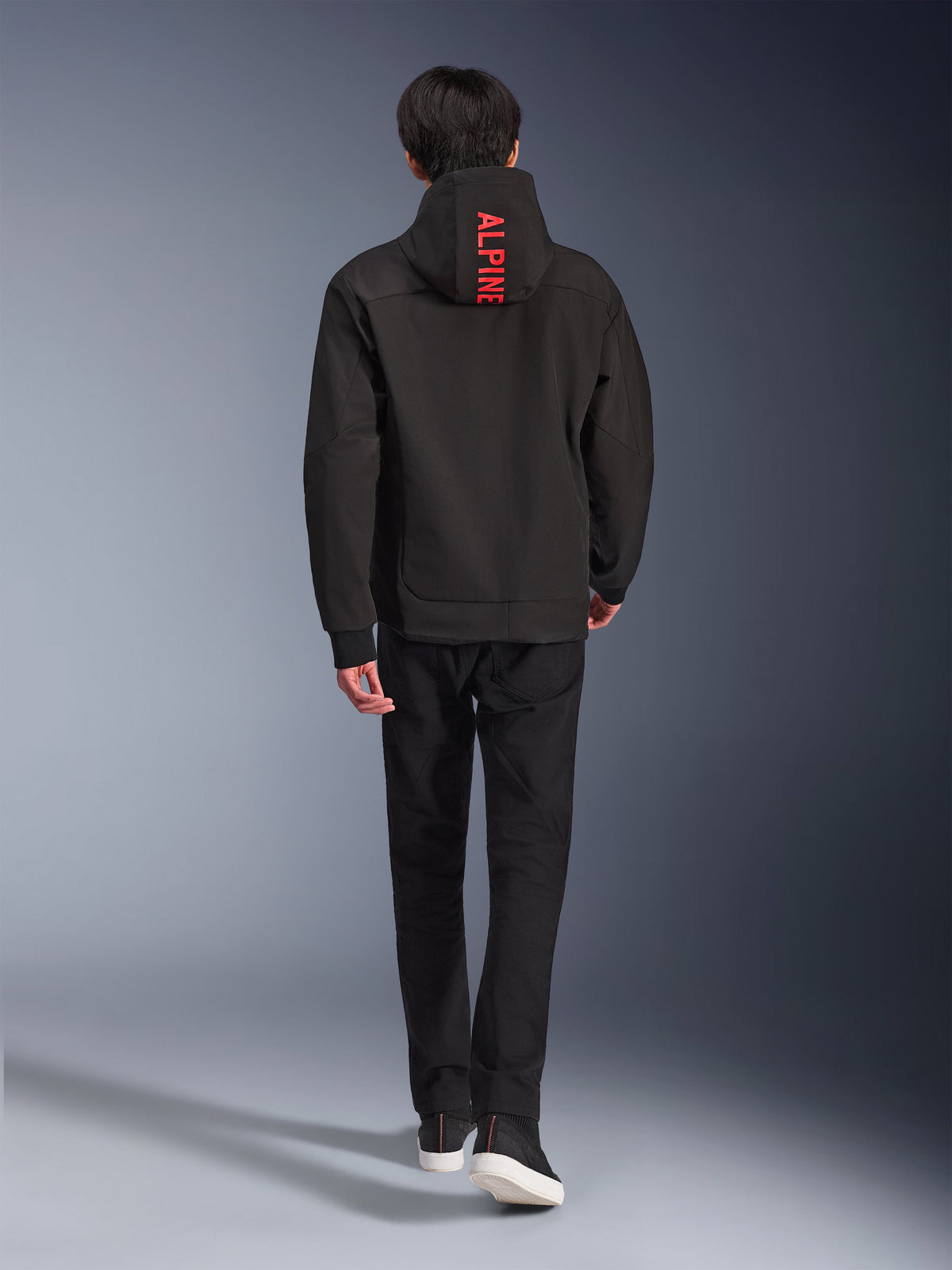 Alpinestars Radium Tech Hoodie, veste de moto urbaine, noir, vue de dos avec le texte « ALPINE » rouge vif sur la capuche, porté avec un pantalon noir et des baskets, coque technique élégante pour les trajets quotidiens.
