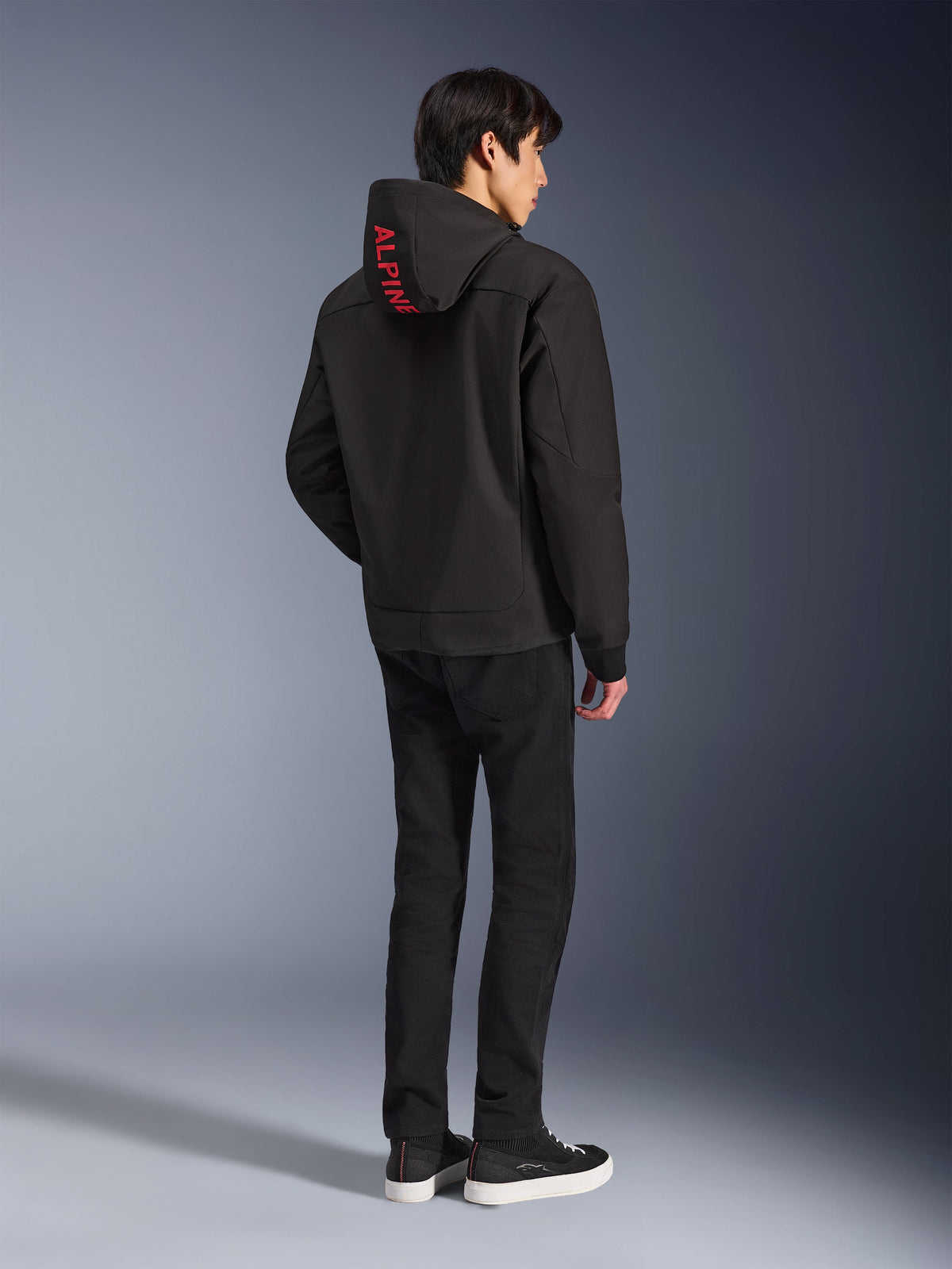 Alpinestars Radium Tech Hoodie, noir avec marquage Alpinestars rouge sur la capuche, vue de dos sur un modèle masculin, porté avec un pantalon noir et des baskets, équipement technique de conduite urbaine